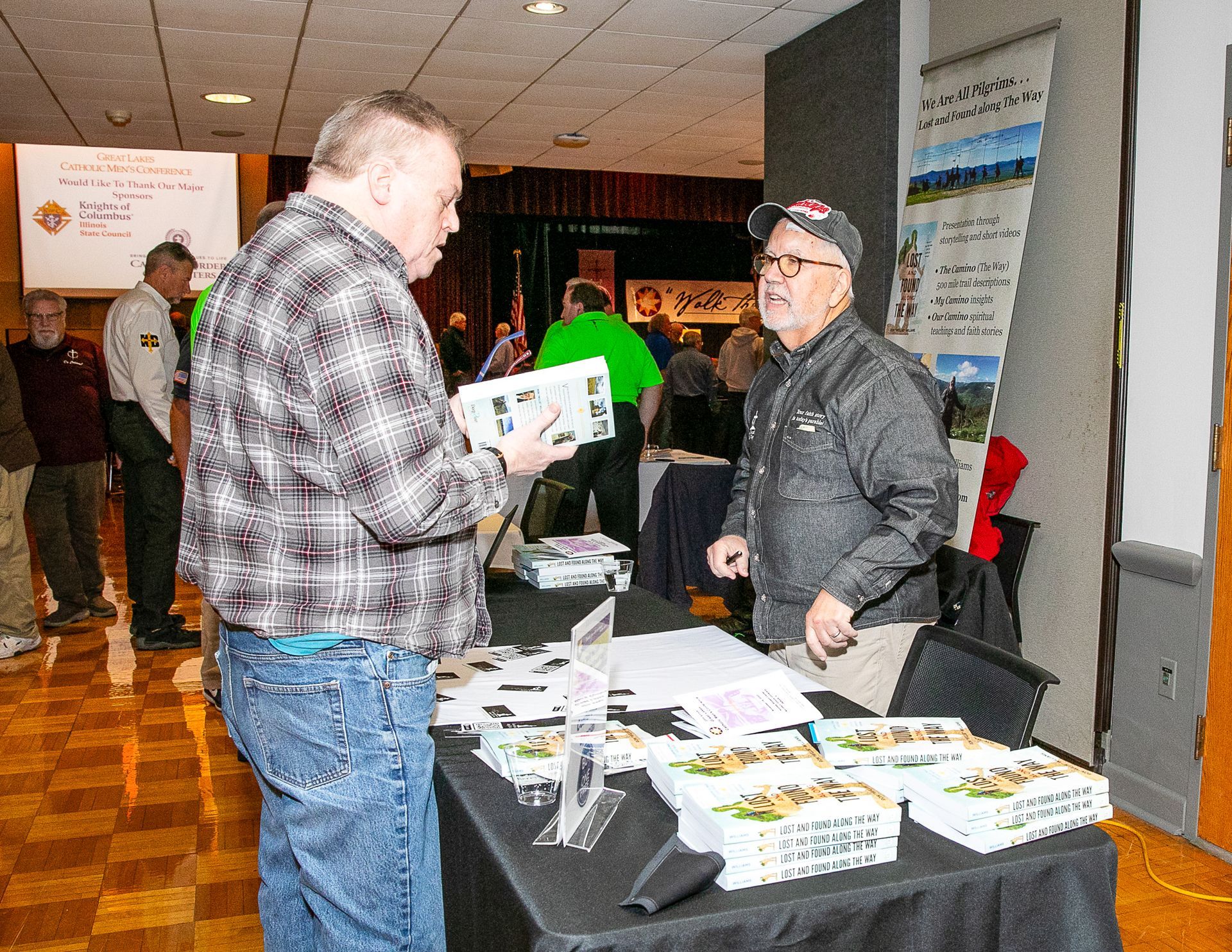 Two people talking at a vendor table with brochures in an indoor event hall.