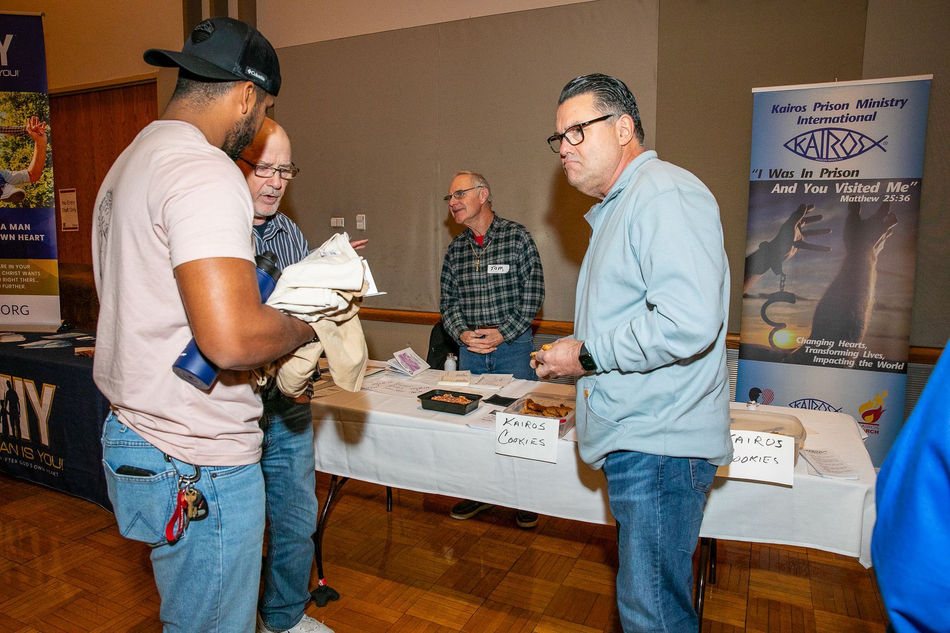 People talking at a booth table during an indoor event, with a banner in the background.