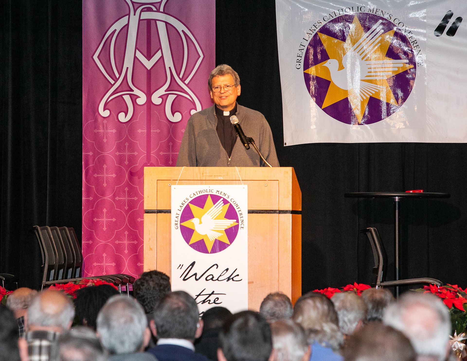 Speaker at podium on stage with purple banners and a crowd in the foreground
