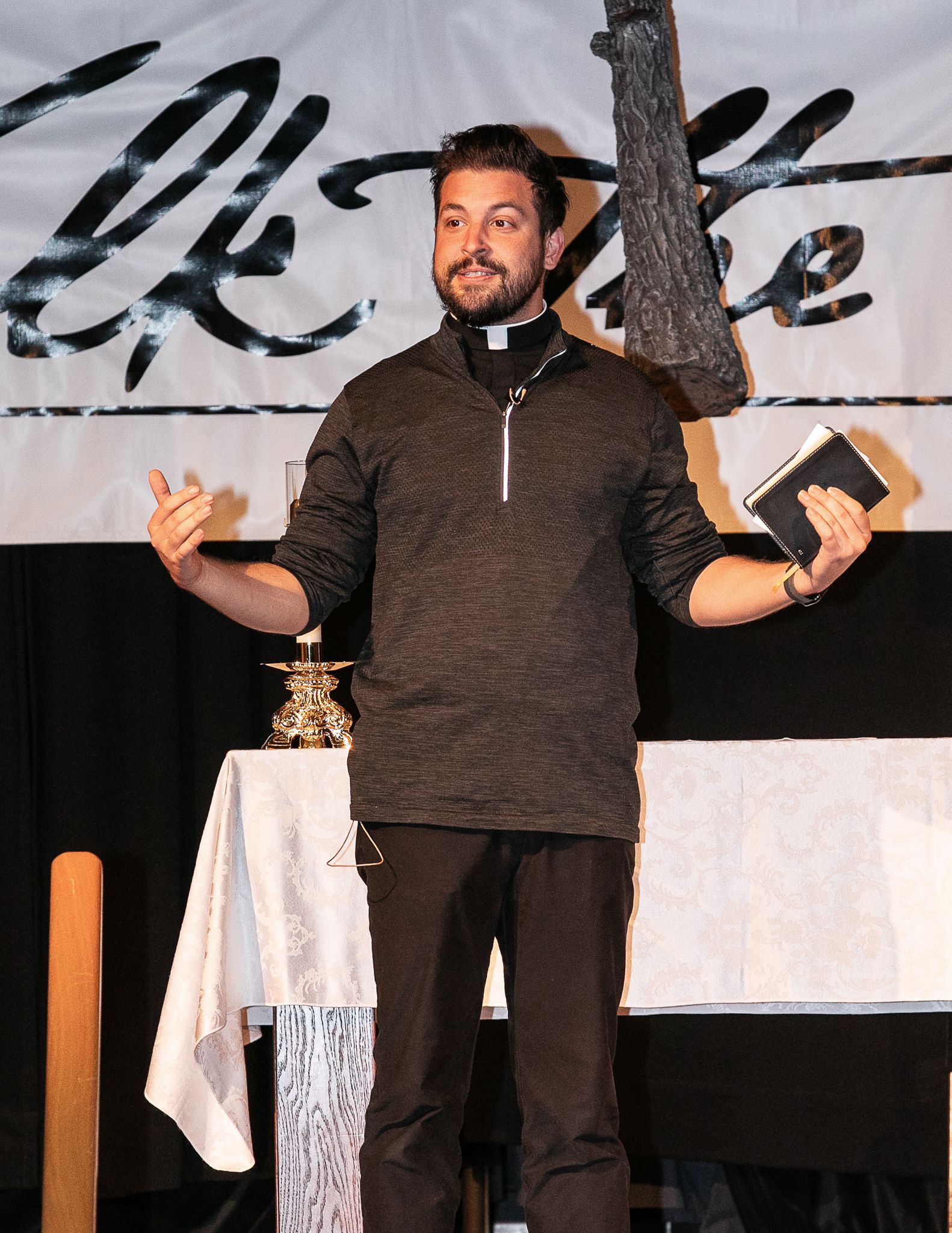 Man standing on stage in a dark pullover, gesturing with both hands before a banner backdrop