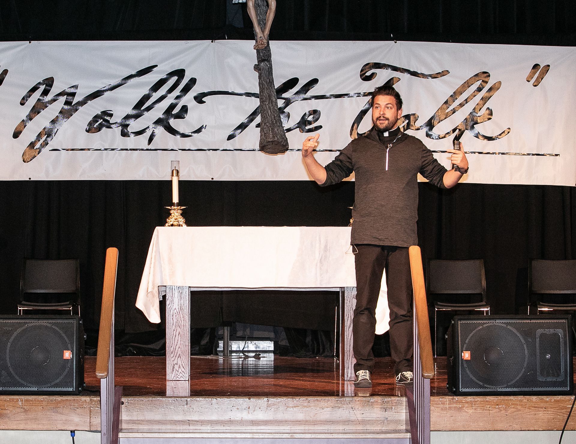 Rev. Timothy Anastos standing on stage beside a table, speaking into a microphone with a banner backdrop.