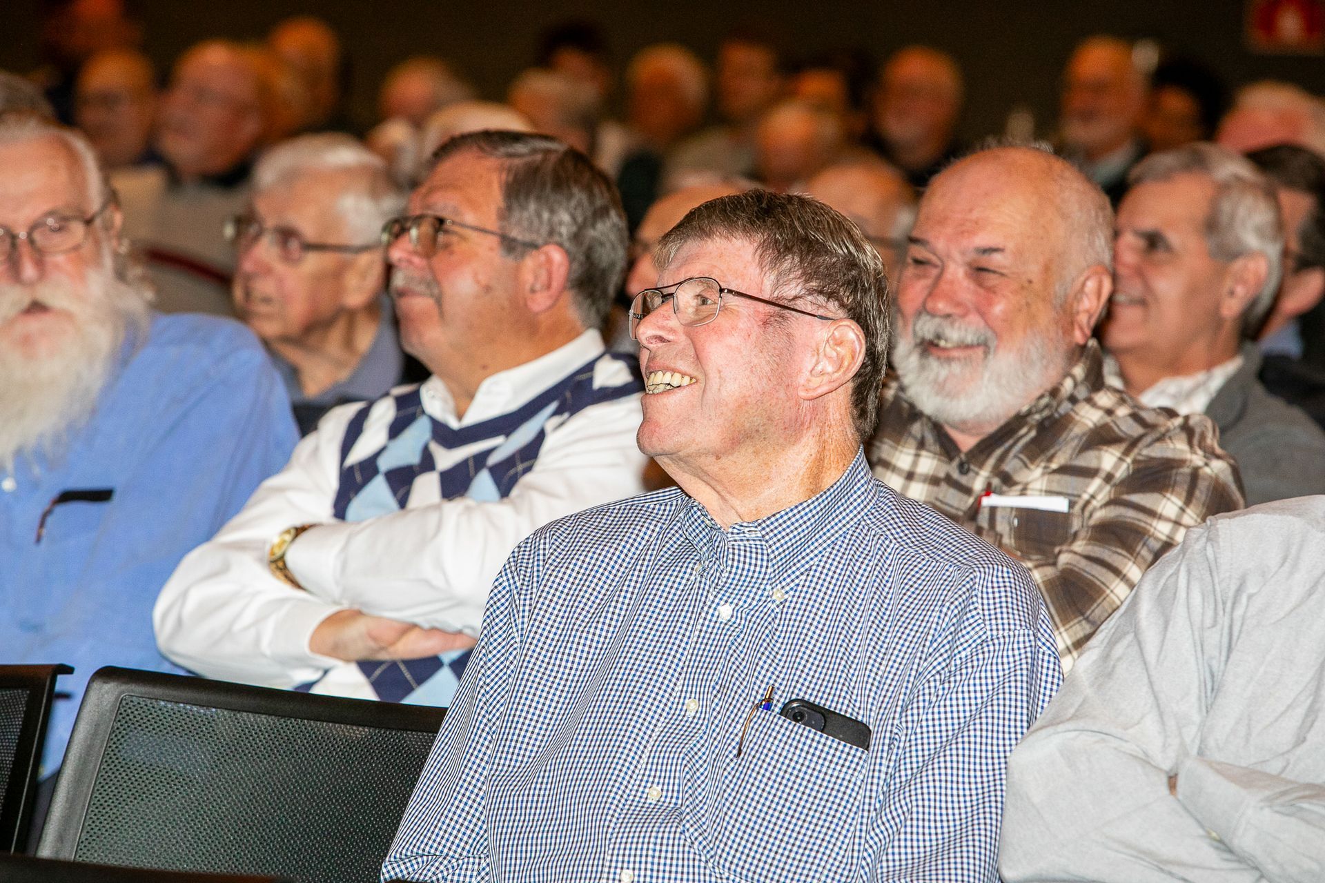 Audience members seated indoors, listening attentively; man in blue checkered shirt in the foreground.
