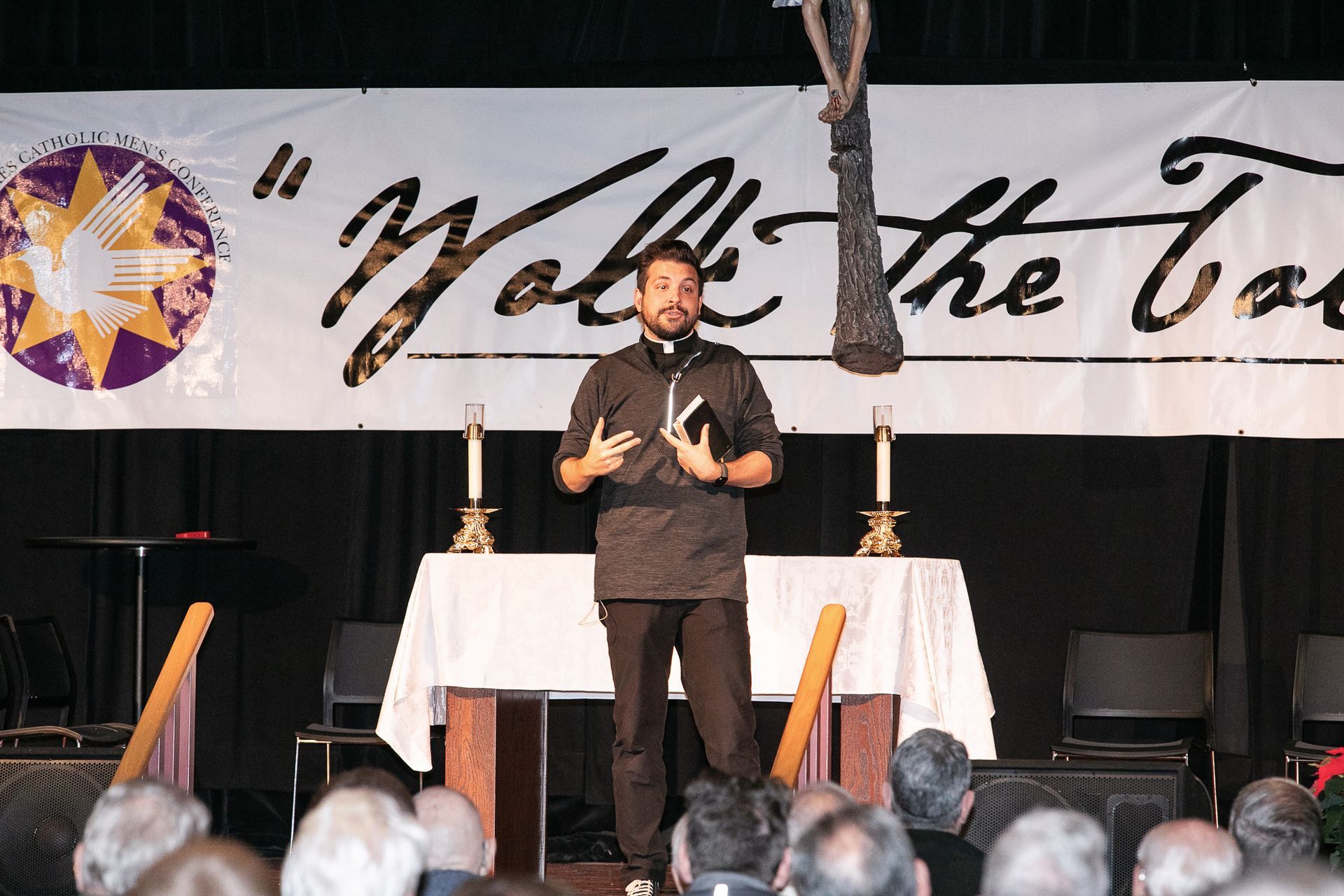 Man speaking onstage before a banner, with audience heads visible in the foreground
