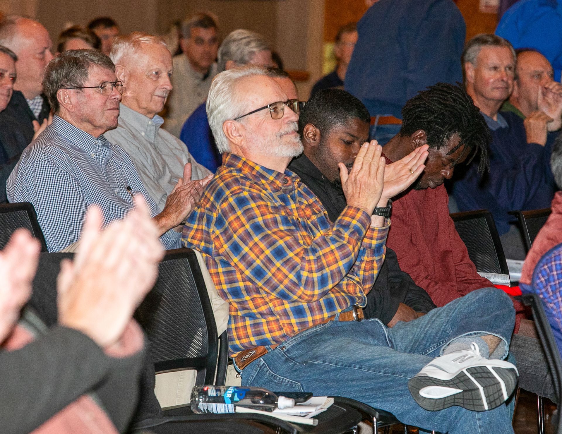Audience seated and applauding at an indoor event, with a man in a plaid shirt in the foreground.
