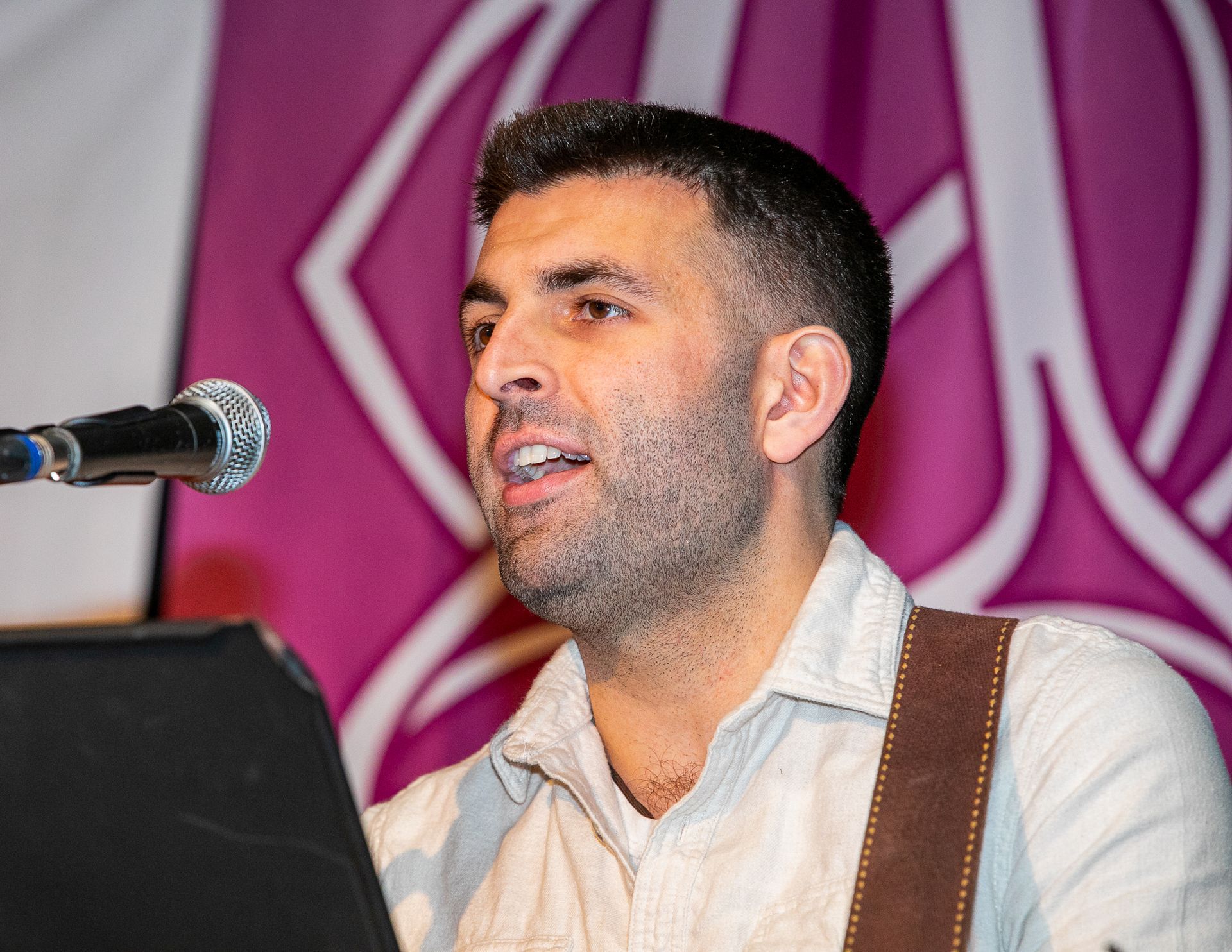 Man speaking into a microphone at a podium, in front of a pink patterned backdrop