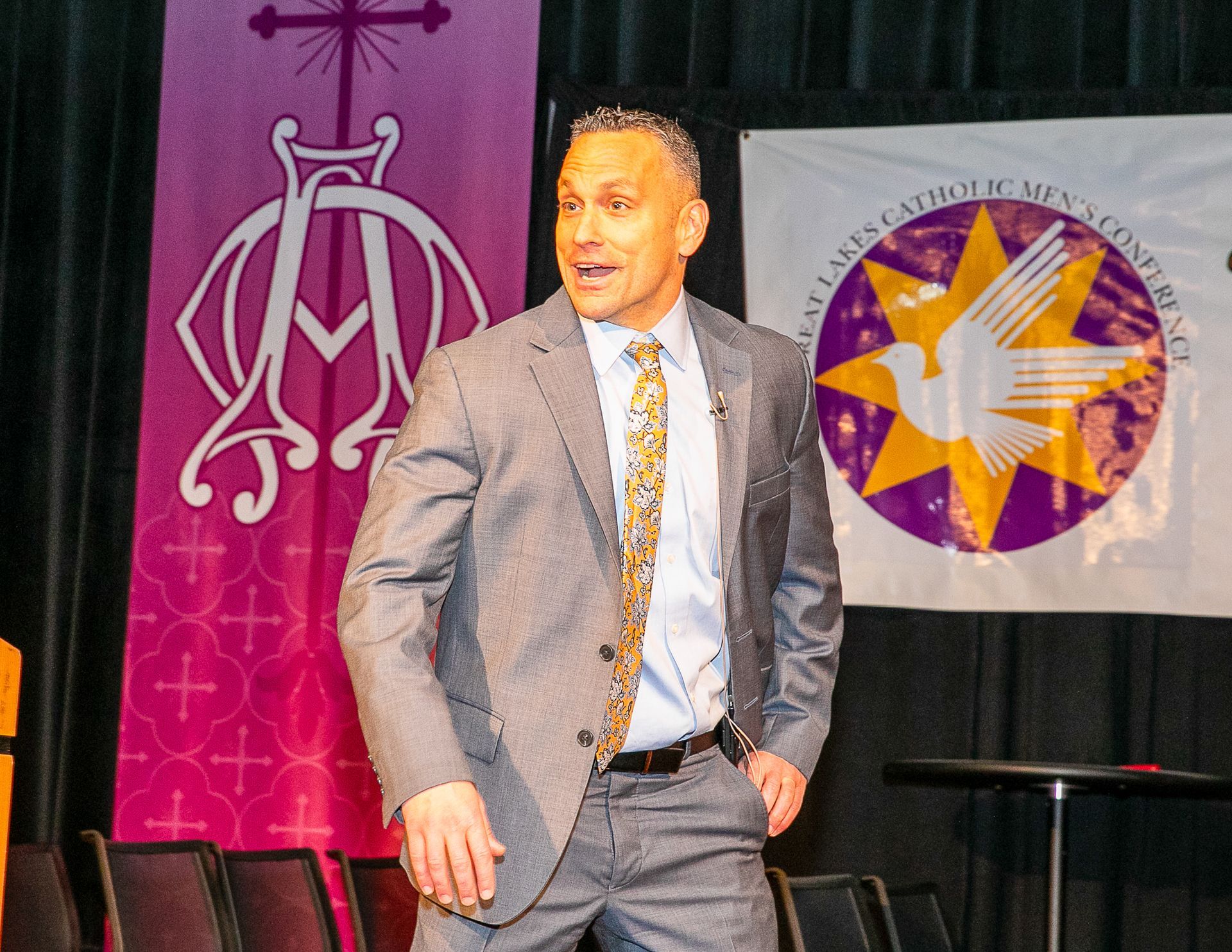 Justin Fatica in gray suit speaking onstage beside banners and a circular emblem backdrop