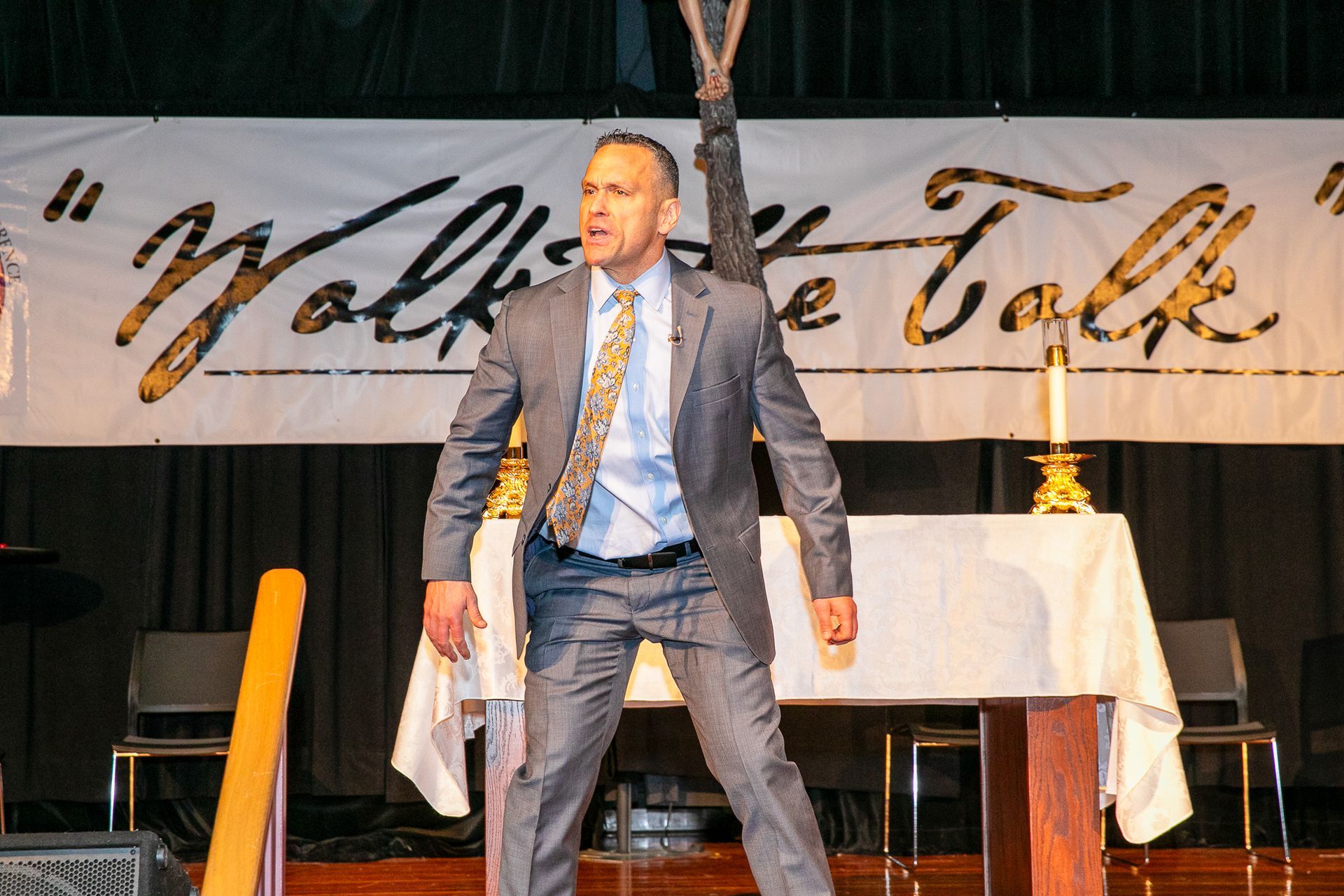 Man in a blue suit speaking on a stage with a table and backdrop banner behind him
