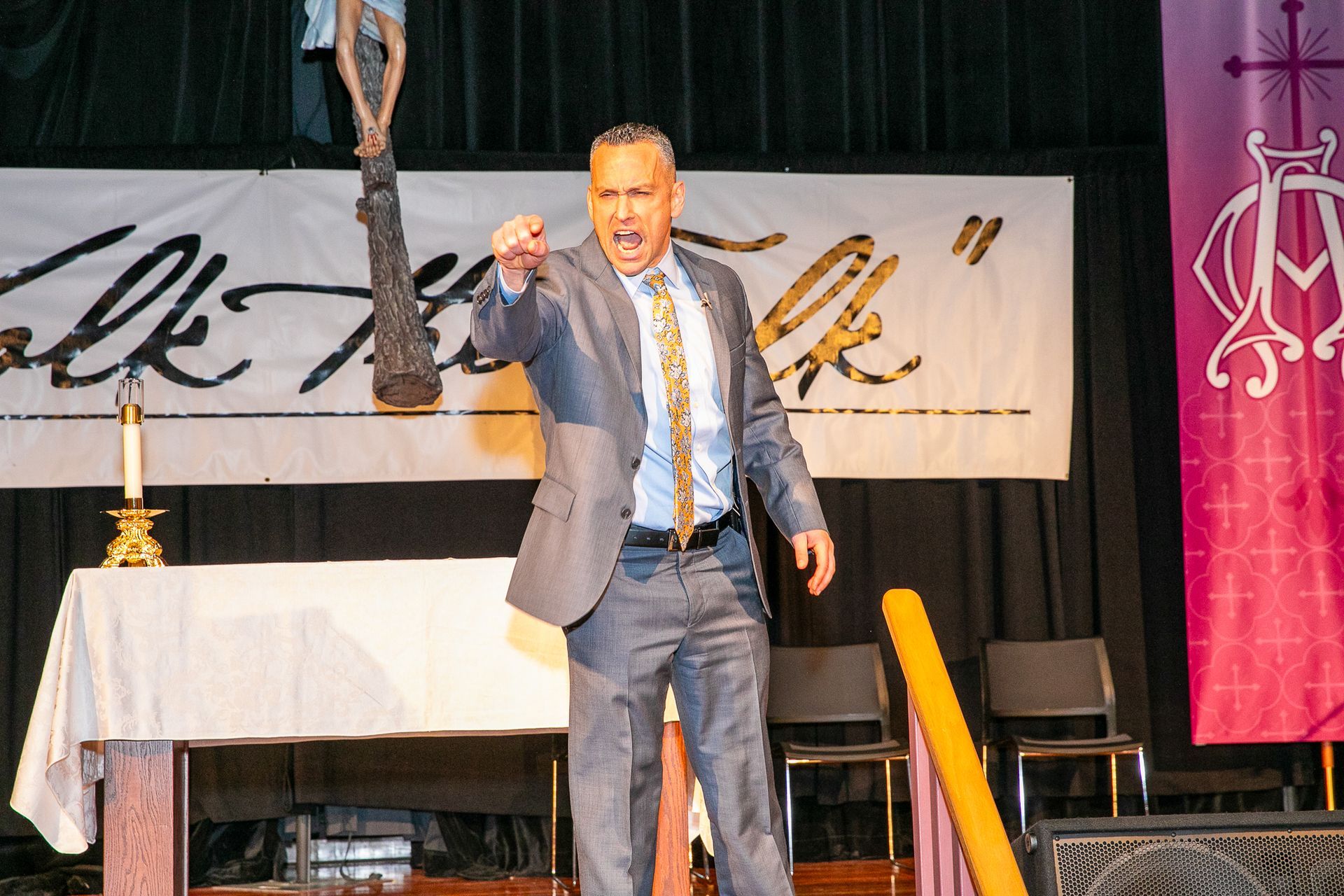 Justin Fatica in suit speaking on stage, pointing forward beside a white table and banner backdrop.