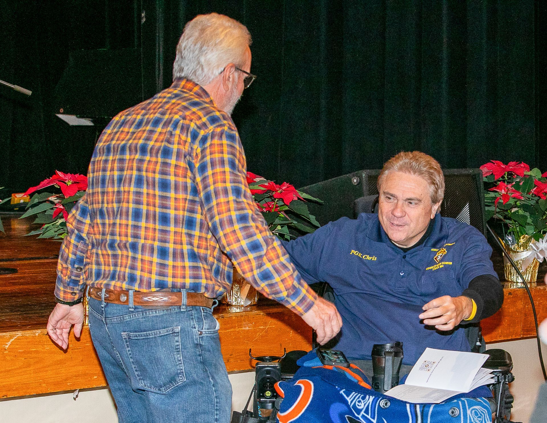 Two men greet at an event, one seated in a wheelchair, with flowers and a stage behind them.