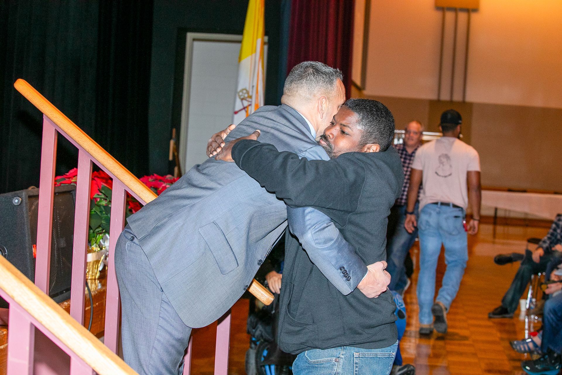 Two people embracing in a hall near a staircase, with another person walking in the background.