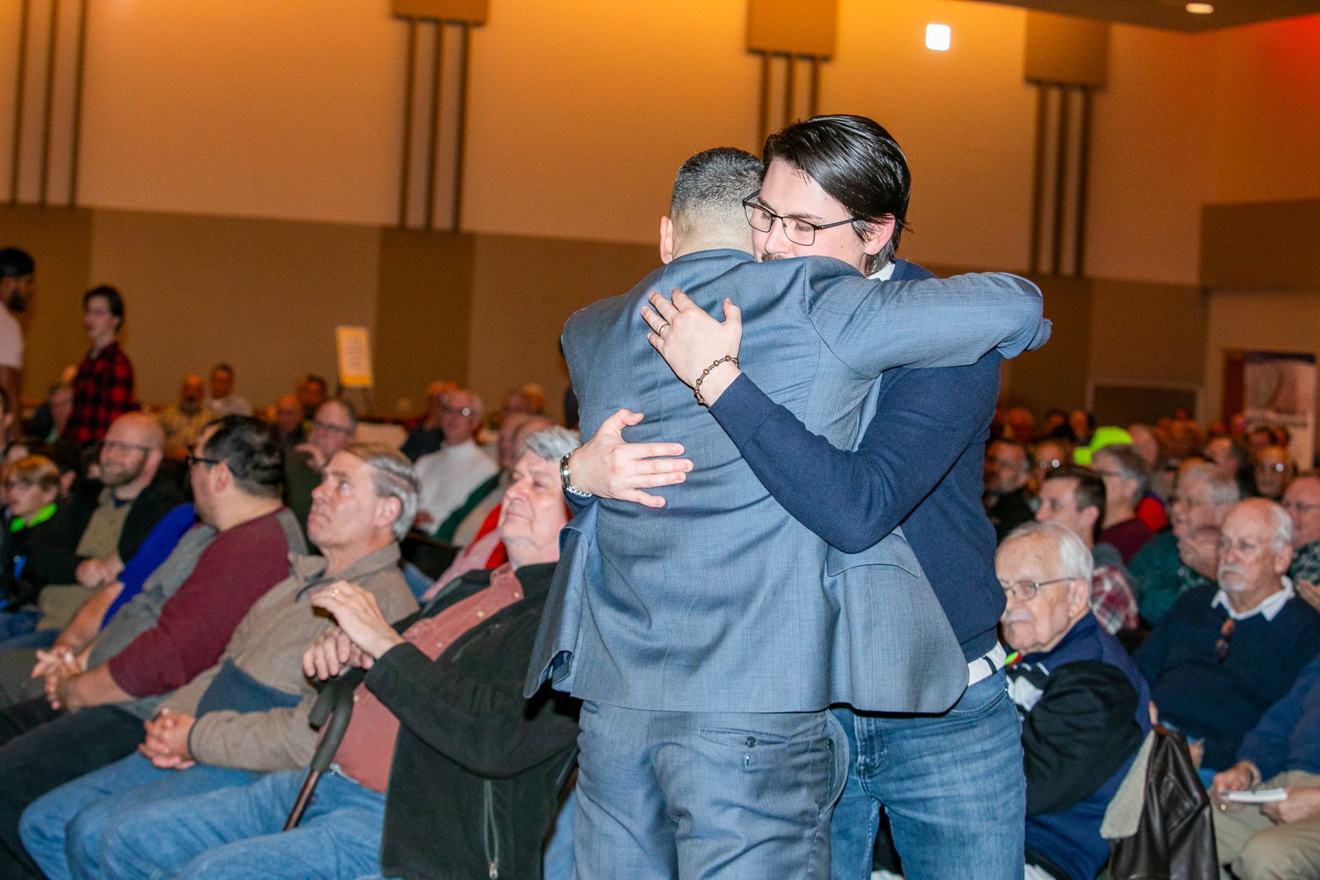 Two people hugging in a crowded auditorium while seated attendees watch nearby