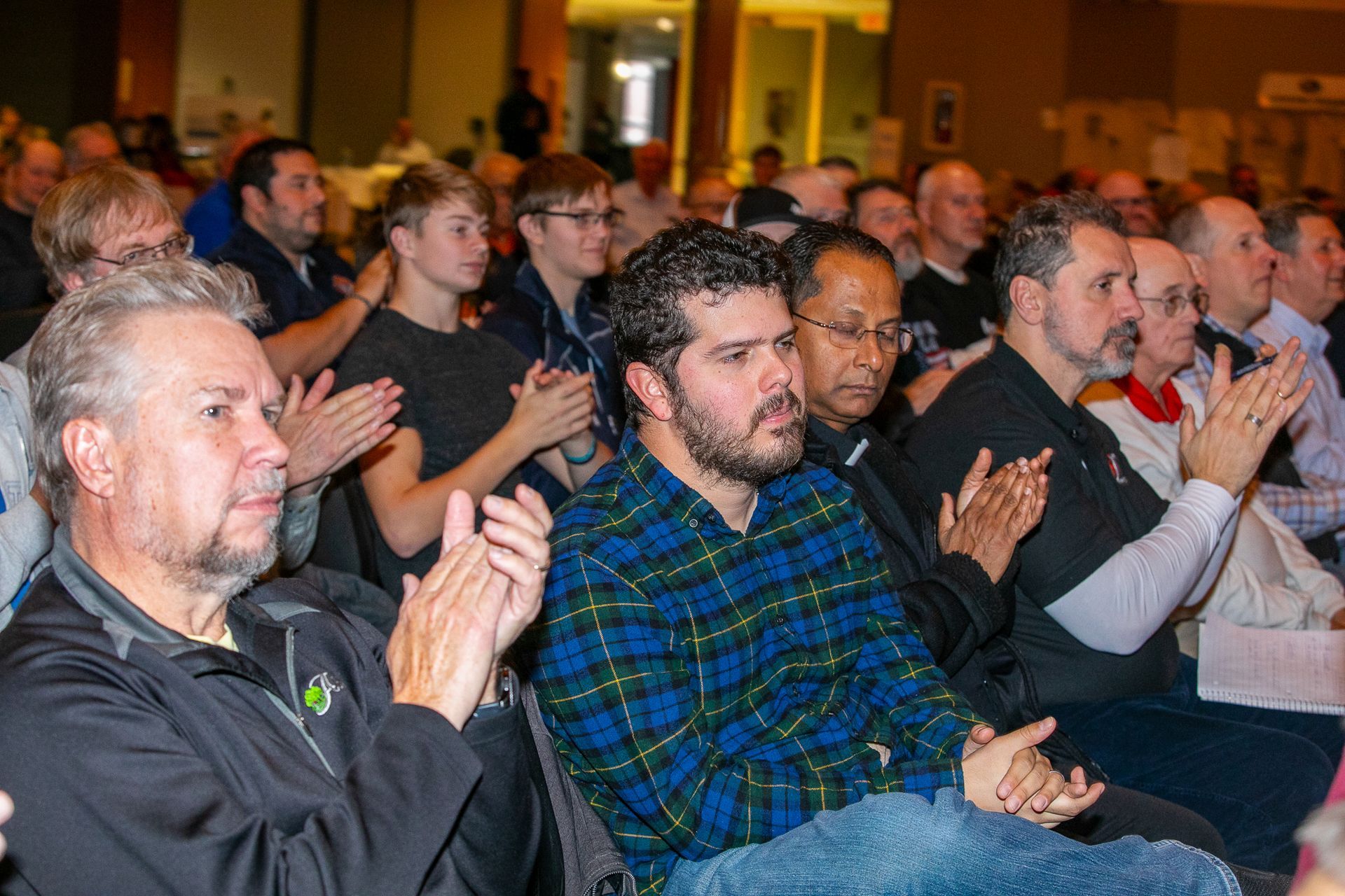Audience seated indoors, applauding at an event, with a man in a blue plaid shirt in the foreground