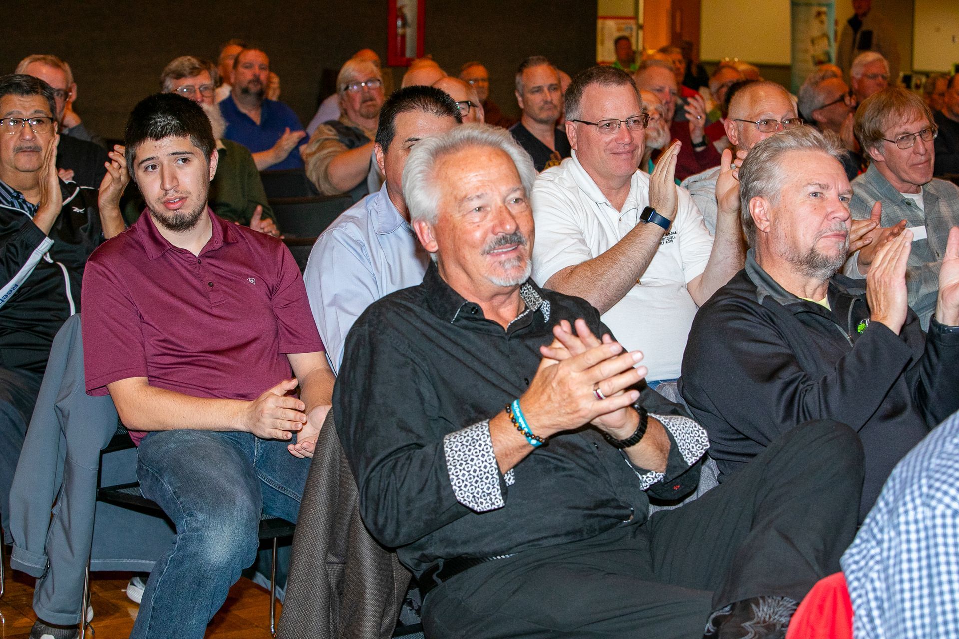 Audience applauding at a seated event, with several people in casual and dark clothing visible.