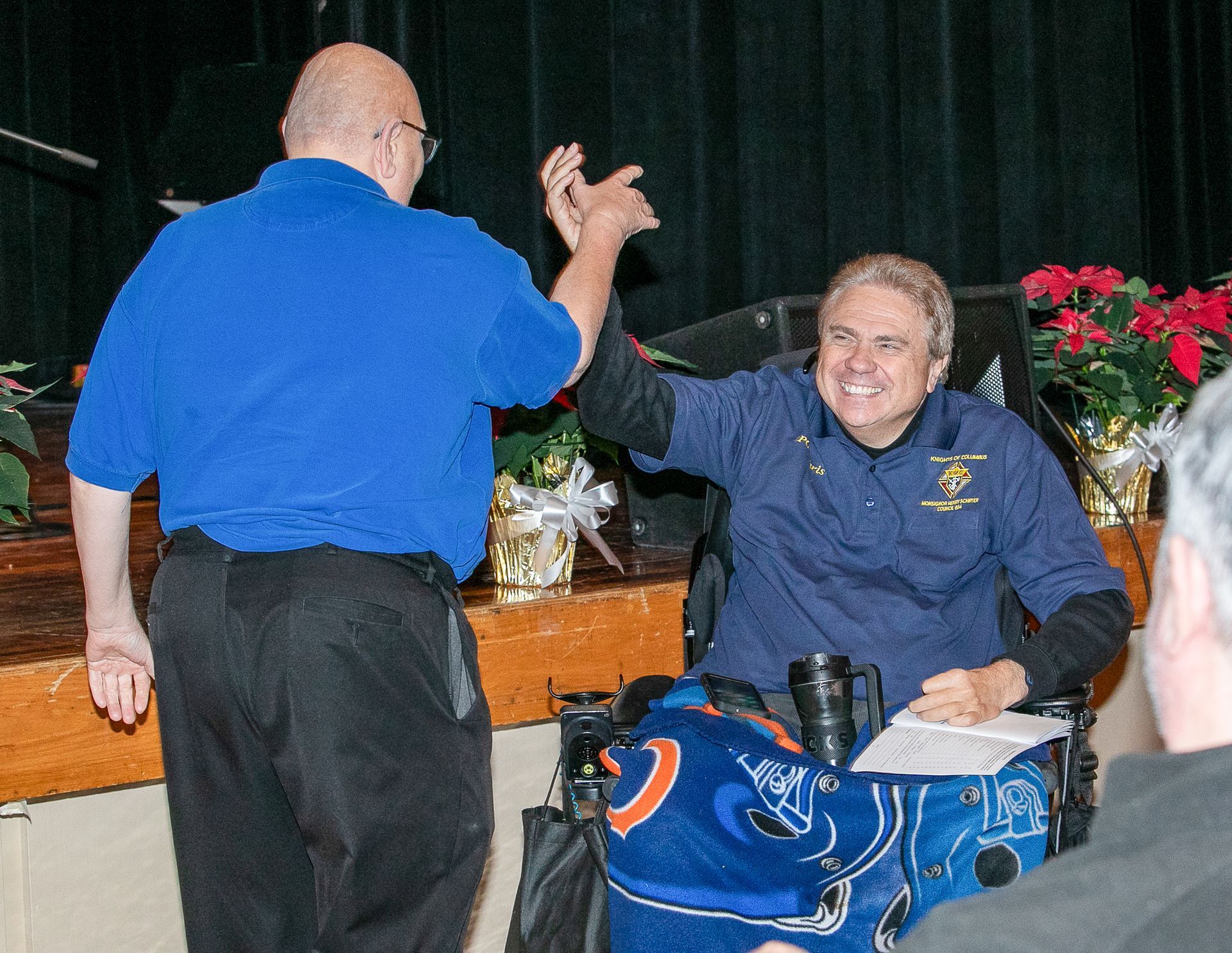 Two men high-five beside a stage, one seated in a wheelchair, with flowers and a blue sports bag nearby.