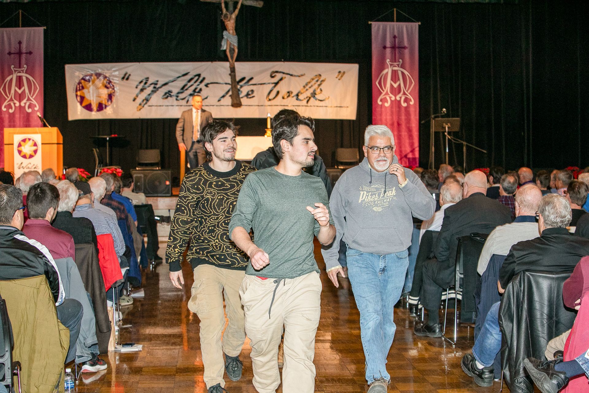 Three people walk through a crowded church hall toward the camera, with seated attendees in the background.