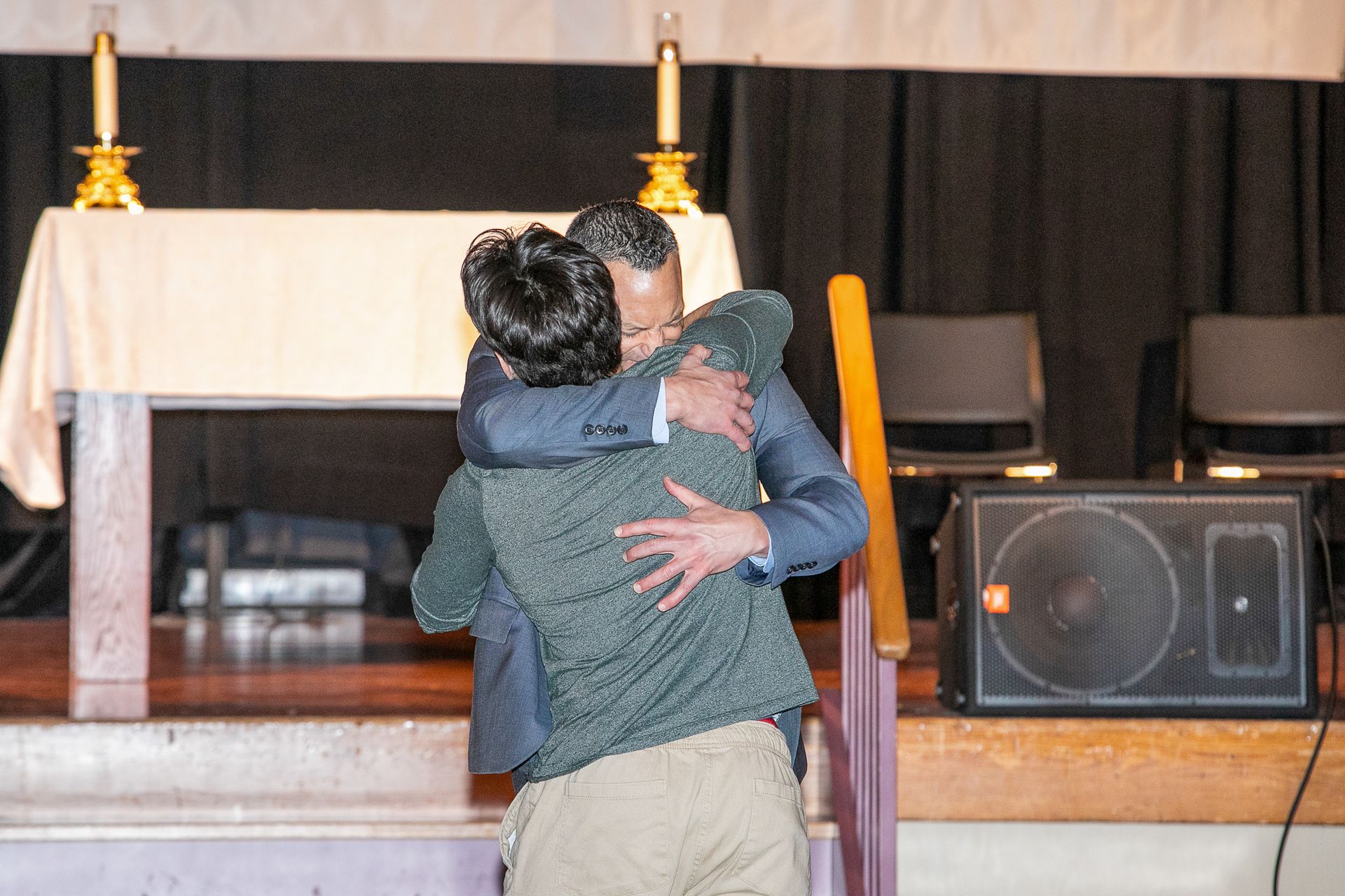 Two people hug on a stage before an altar with candles and speakers in the background.