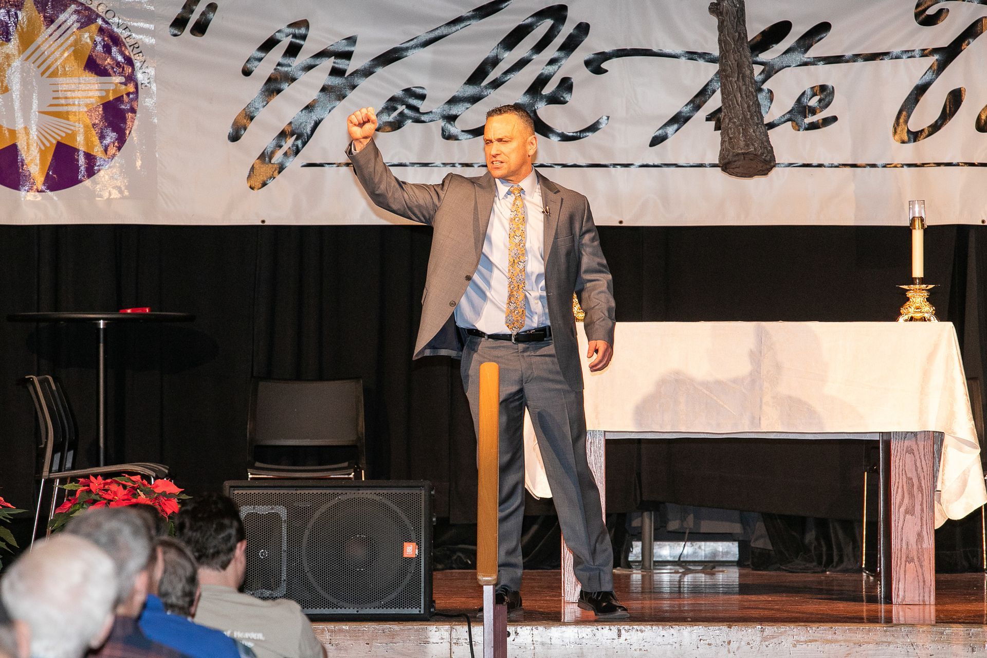 Justin Fatica on stage raising one hand beside a table and microphone at a Catholic men's conference.