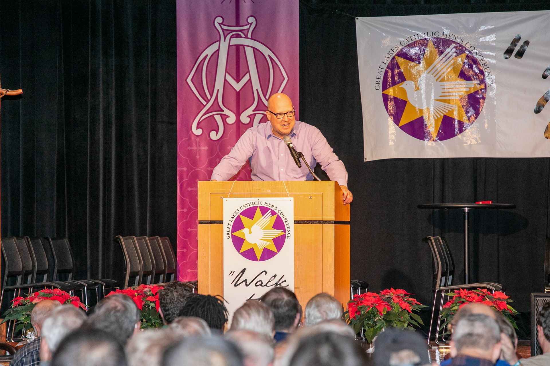 Man speaking at a podium on a stage with maroon and white banners, audience in foreground