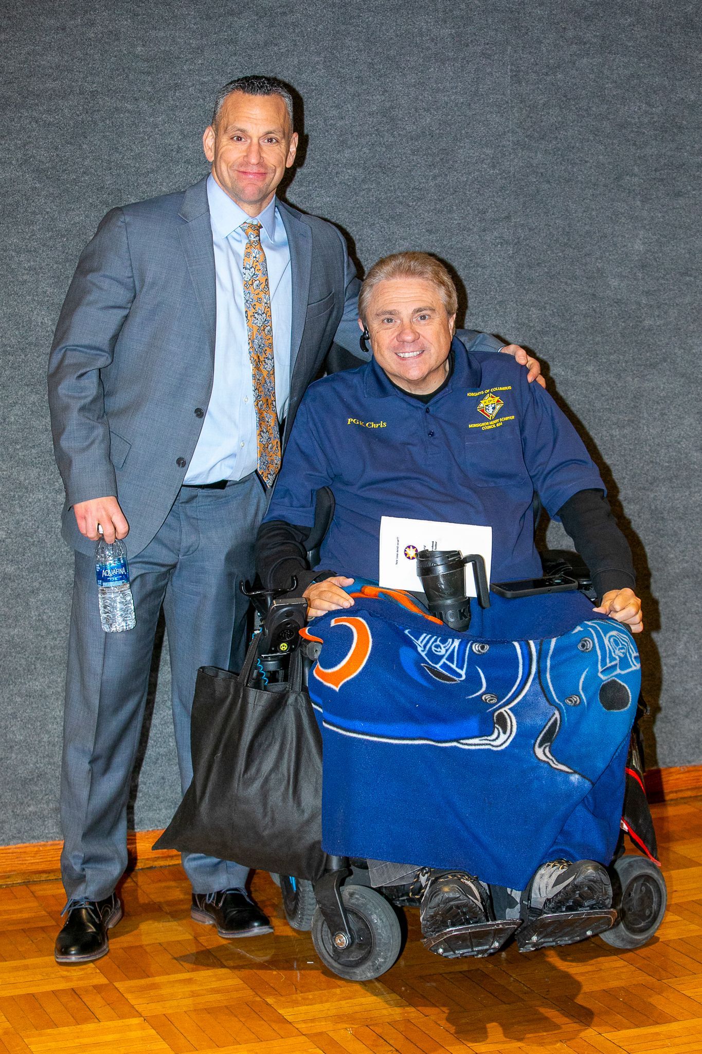 Two men pose indoors; one stands beside a man in a wheelchair wearing a blue sports blanket.