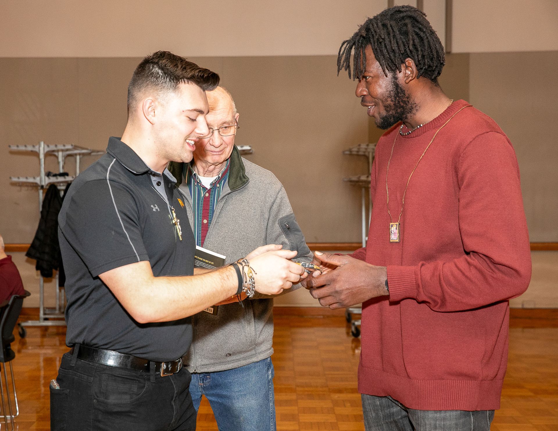 Three people exchanging a small item in a wood-floored room, smiling and talking.