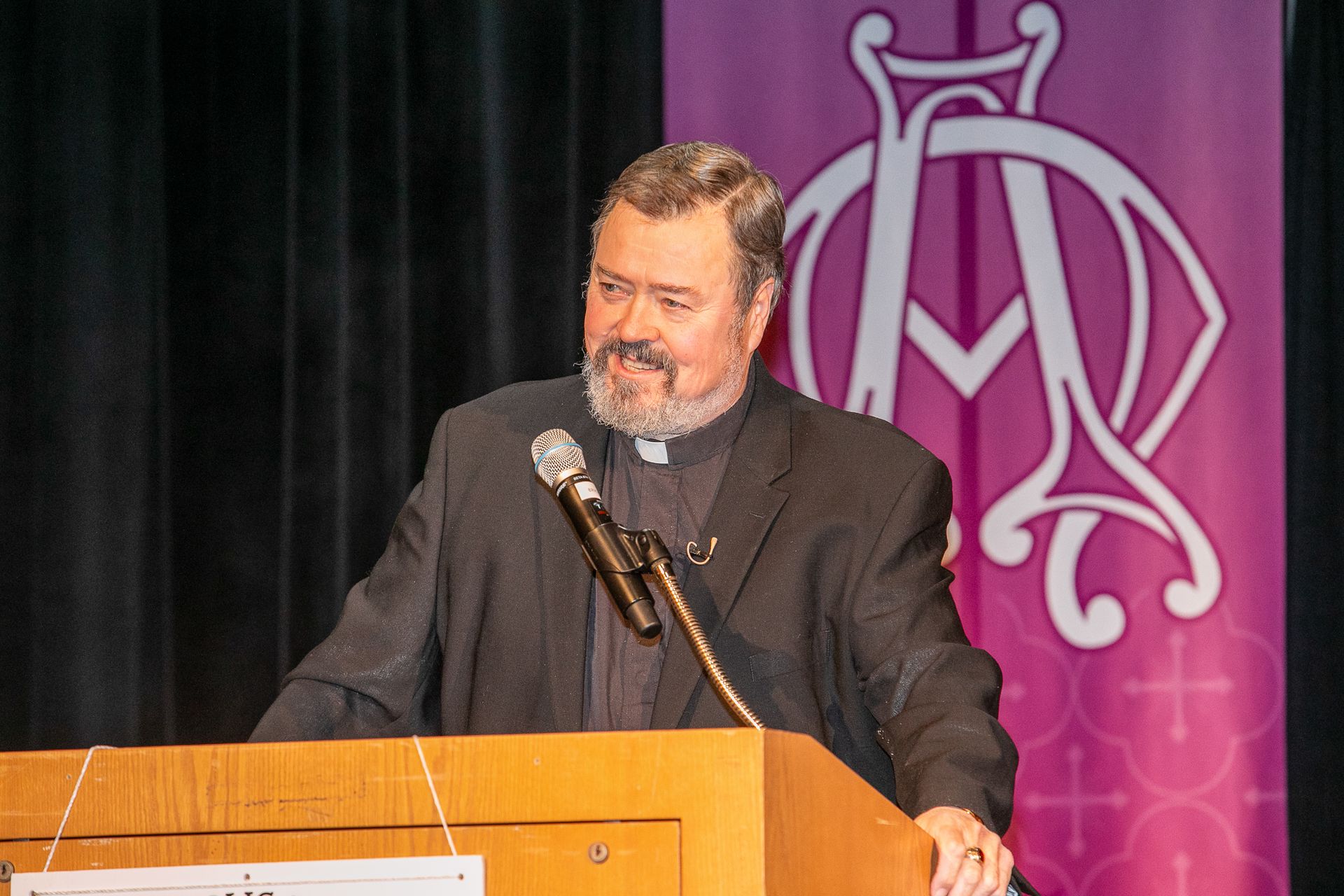 Rev. Richard Simon speaking at a podium on a stage at a Catholic men's conference.