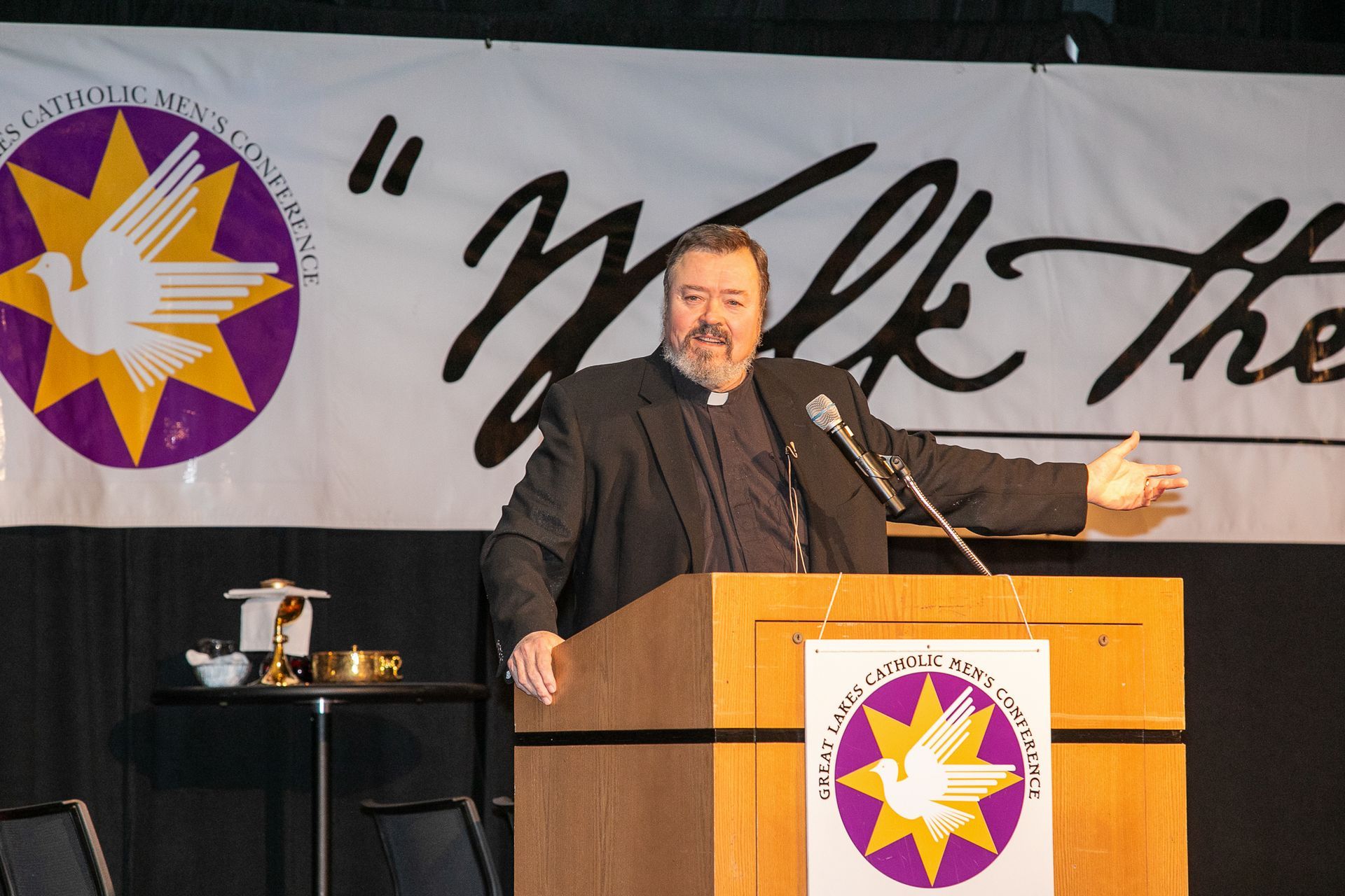 Rev. Richard Simon speaking at a podium during a Catholic men's conference, with a banner backdrop and logo behind him.