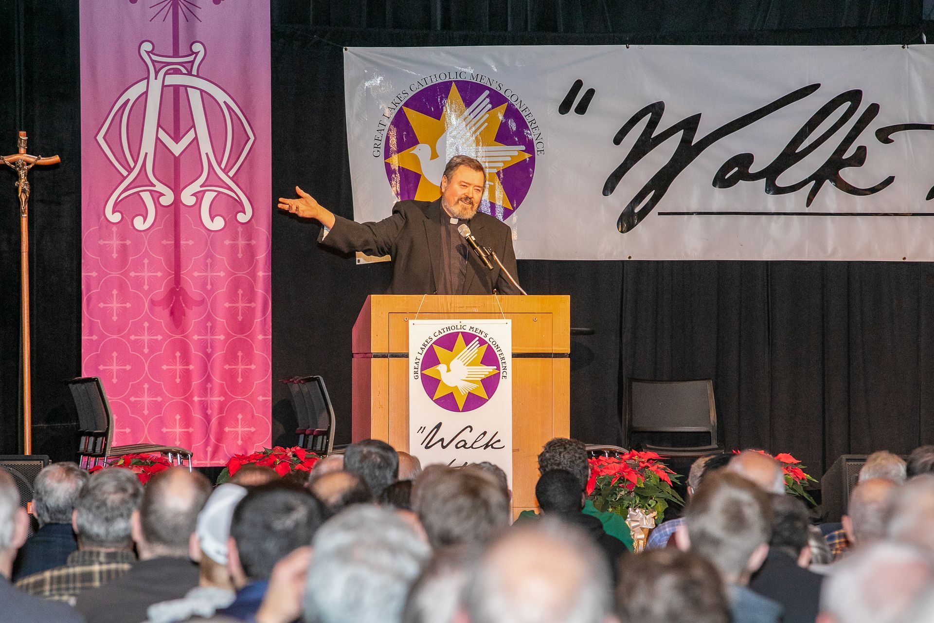 Speaker at podium with raised hand on stage, audience in foreground, pink banner and “Wolf” sign behind