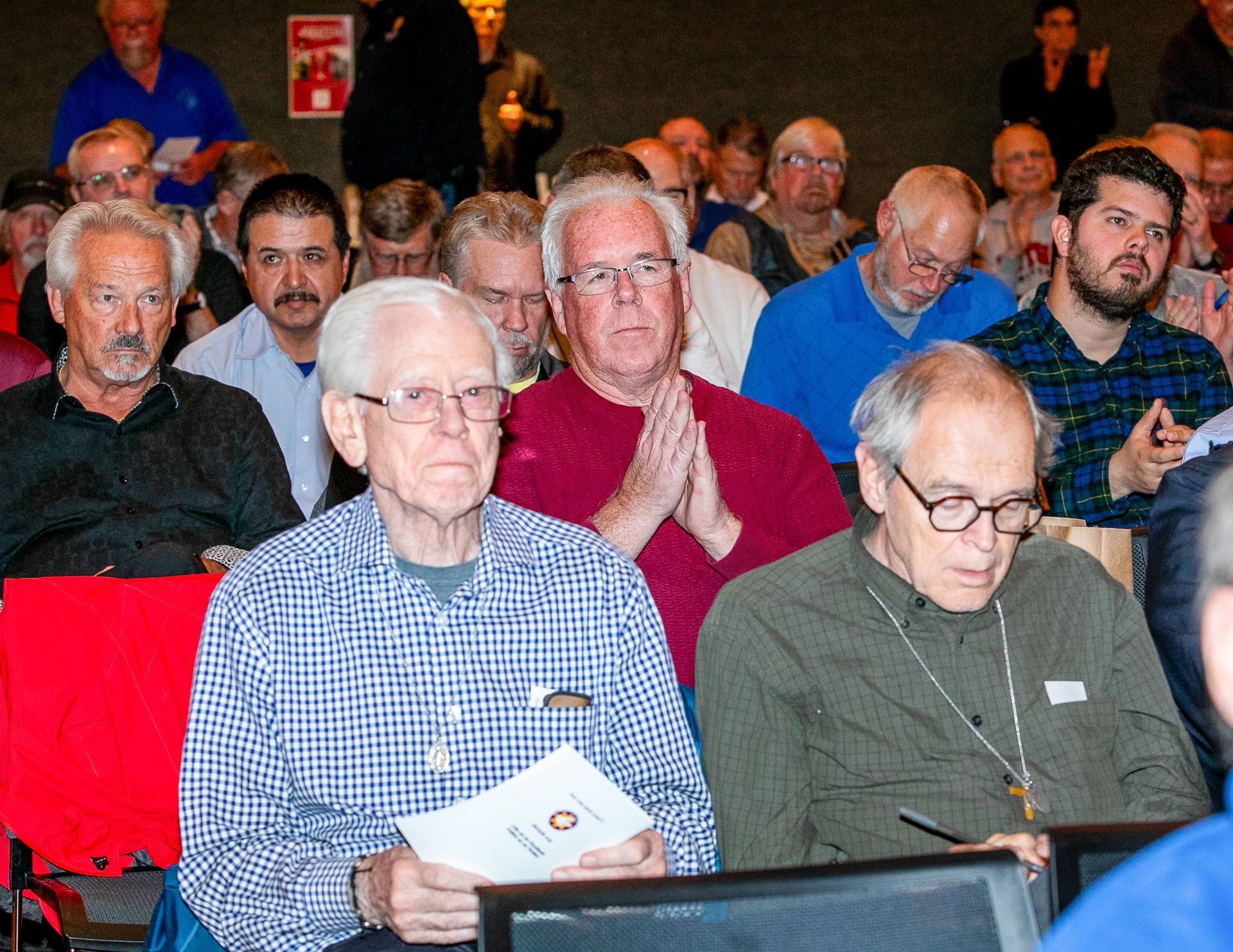 Audience seated indoors, with one man in front holding papers and another looking down at a laptop.