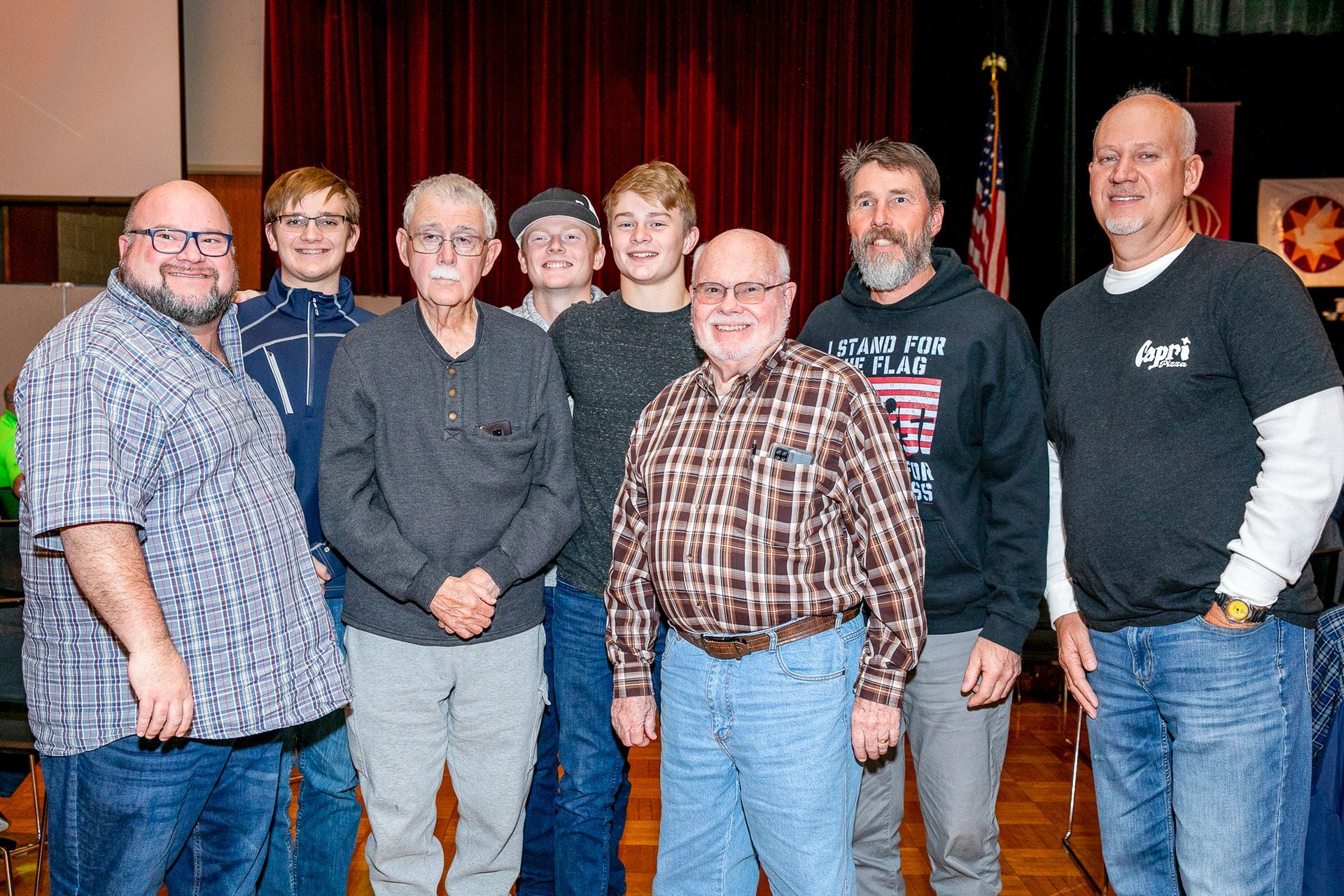 Seven people standing together on a stage in front of red curtains, posing for a group photo.