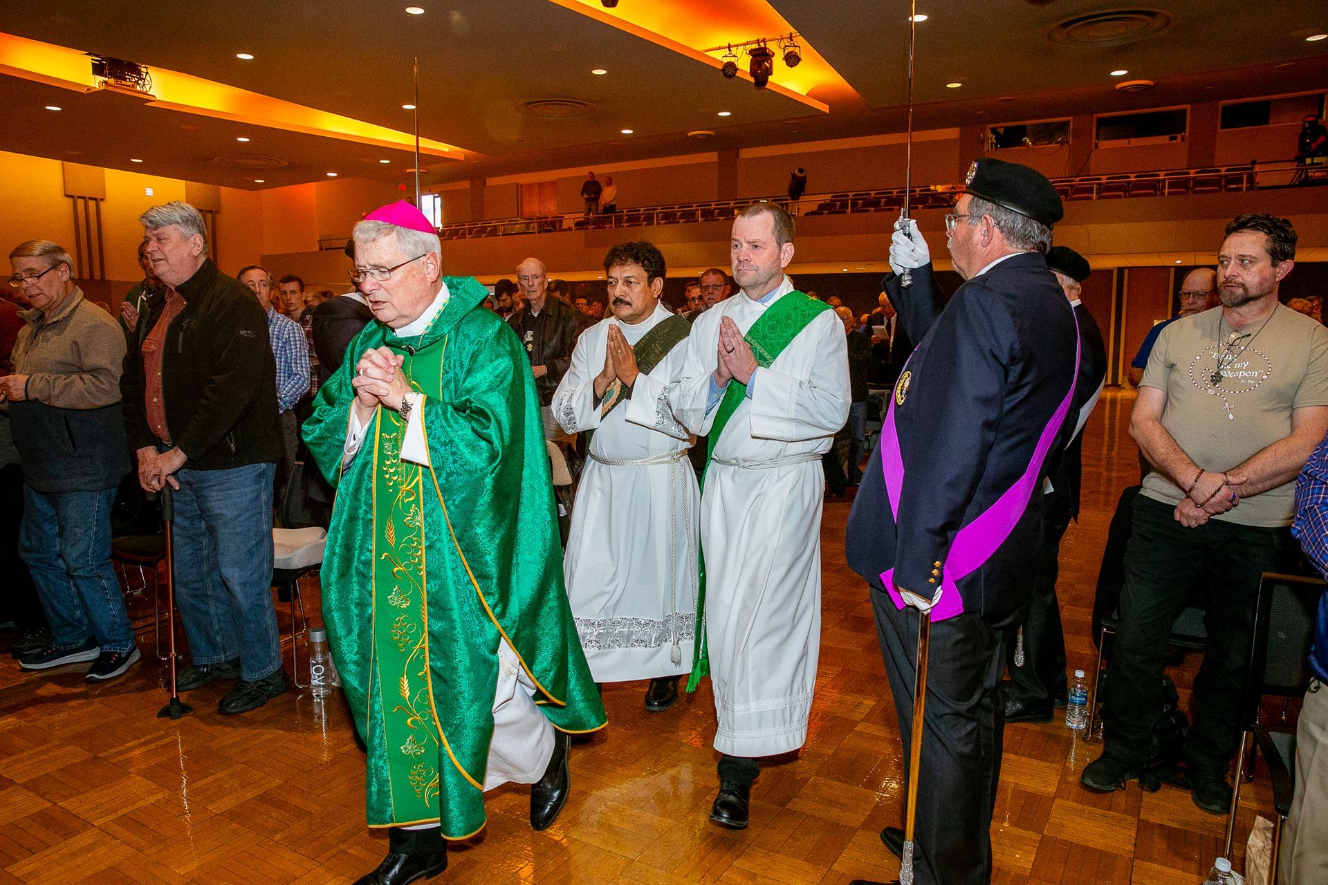 Clergy and Knights of Columbus standing together in a warmly lit hall, one holding a microphone.