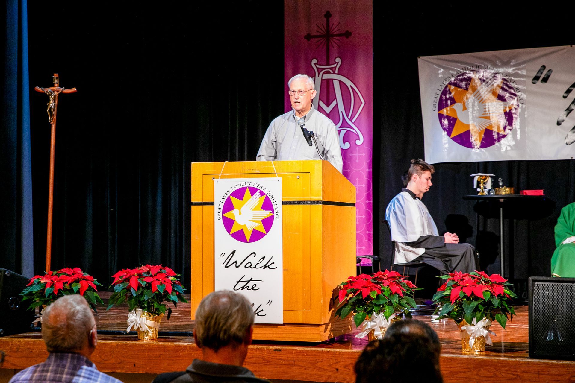 Speaker at podium during church event, with pianist and floral decorations on stage