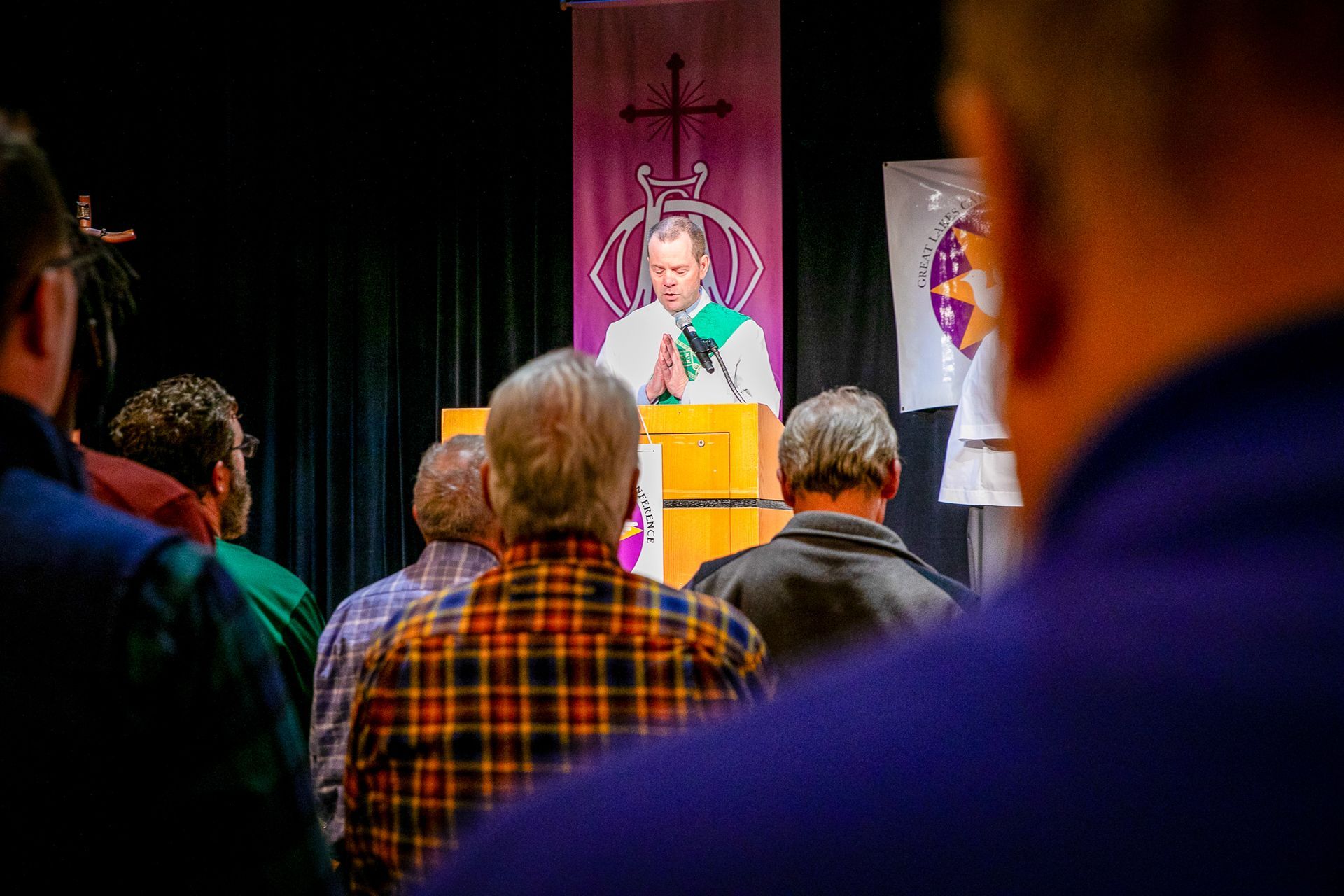 Audience watching a speaker at a podium in a church or chapel, with a cross and stained-glass backdrop
