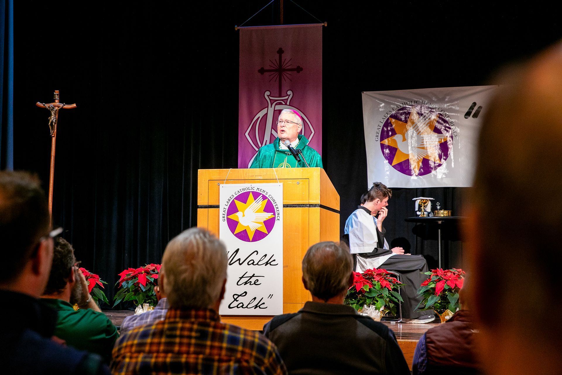 Bishop Malloy at podium on stage during church service, with cross, banners, and seated audience in foreground