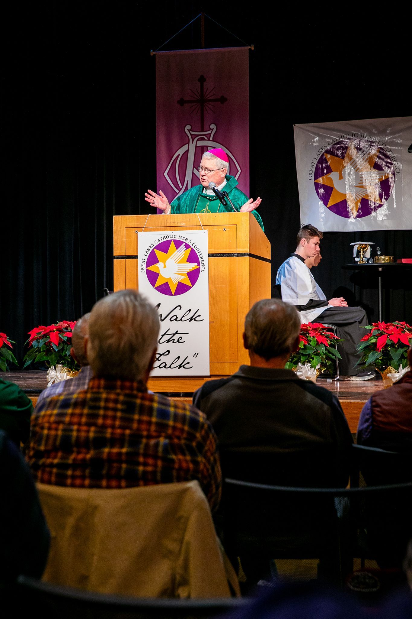 Speaker at podium on stage with seated pianist and audience in foreground, decorated with banners and poinsettias