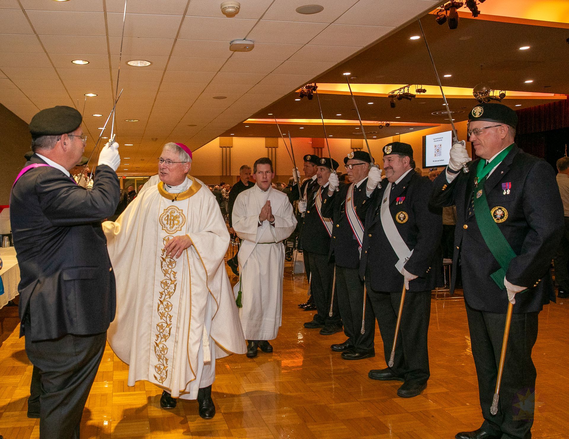 Clergy and suited men stand in a banquet hall during a formal procession.