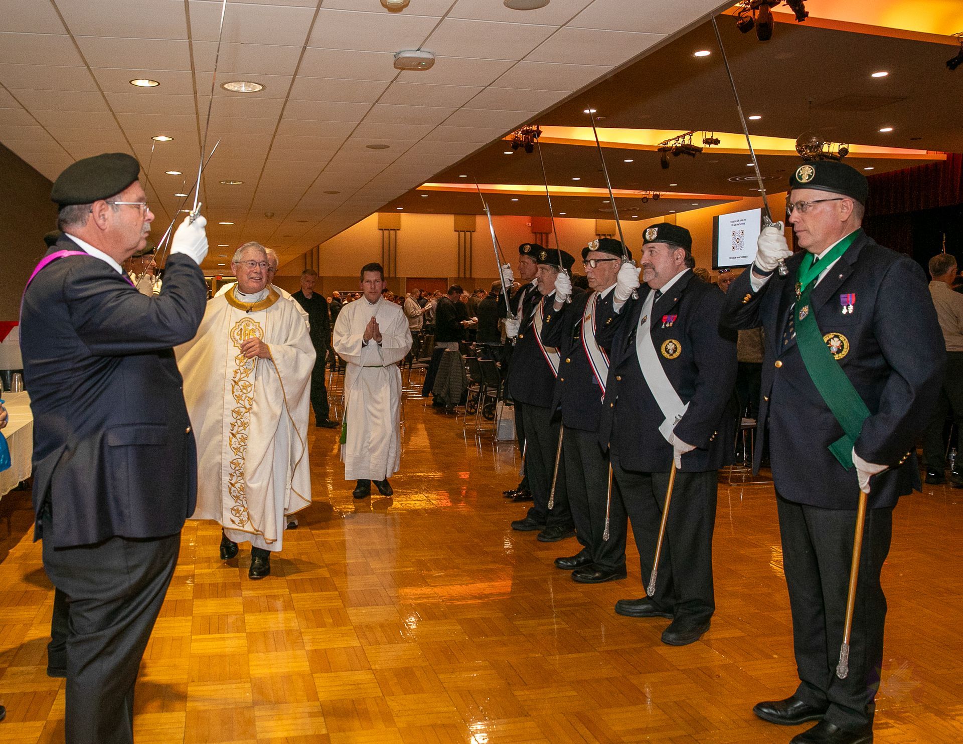 Clergy procession in a banquet hall, with a man saluting on the left and suited attendees lined up on the right.