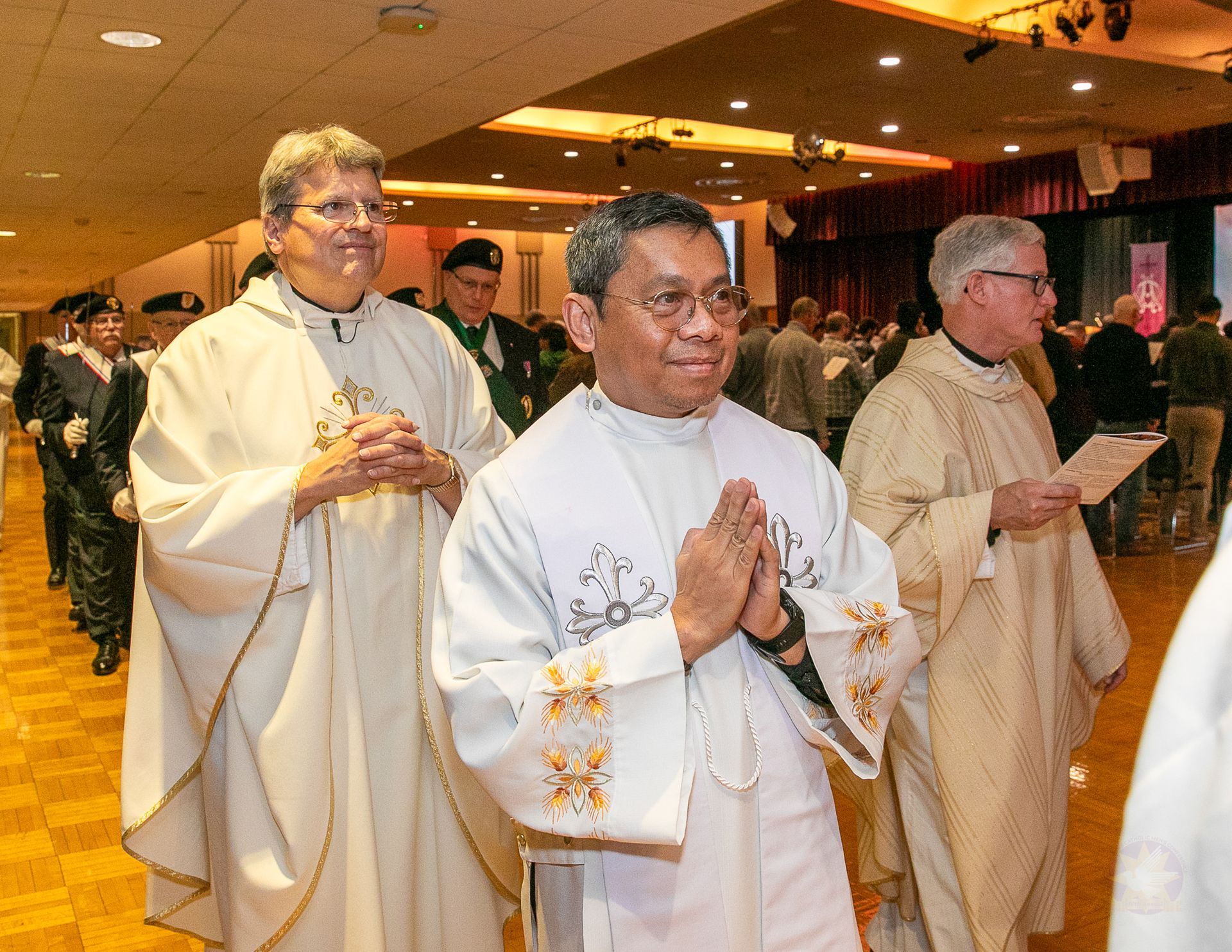 Clergy in white robes walk in a church procession, hands clasped in prayer.