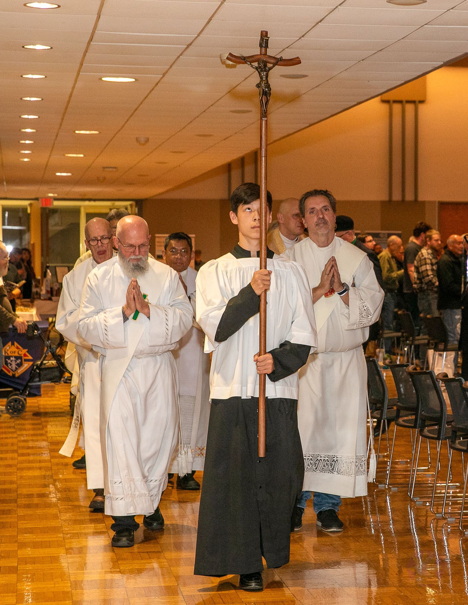 Religious procession indoors, with a crucifix carried by a robed leader followed by clergy in white robes.
