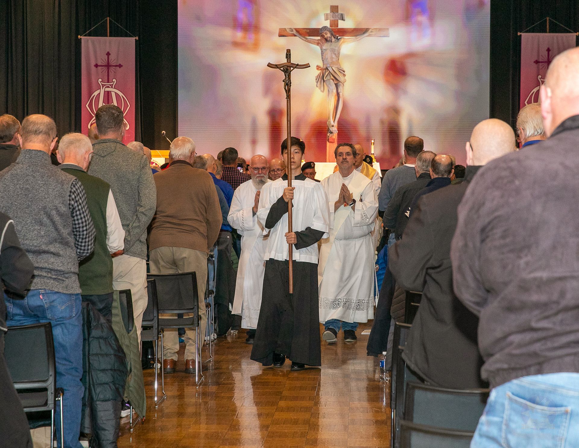 Church procession with clergy holding a crucifix, viewed from the back of a crowded aisle