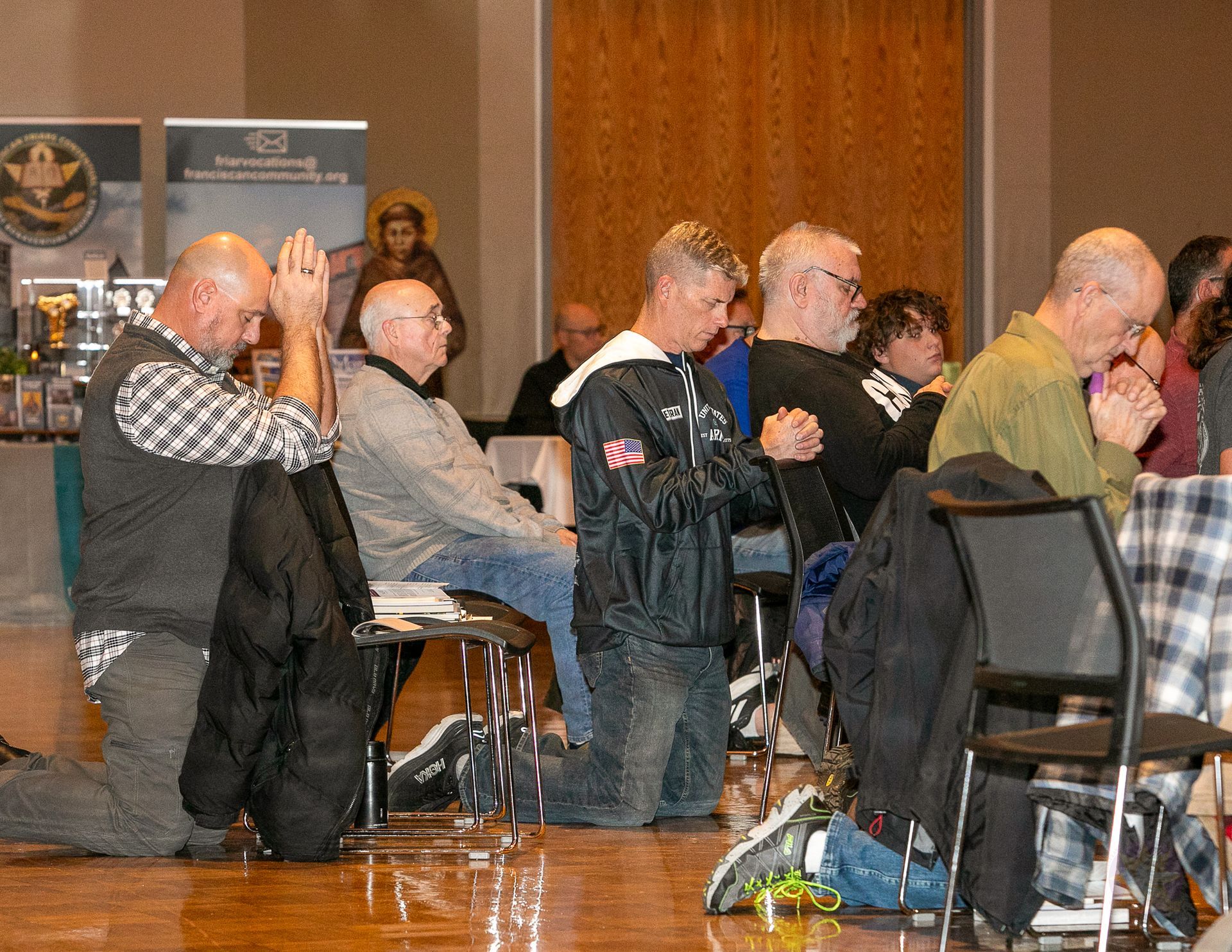 Audience seated in a hall, many praying with folded hands at a Catholic men's conference.