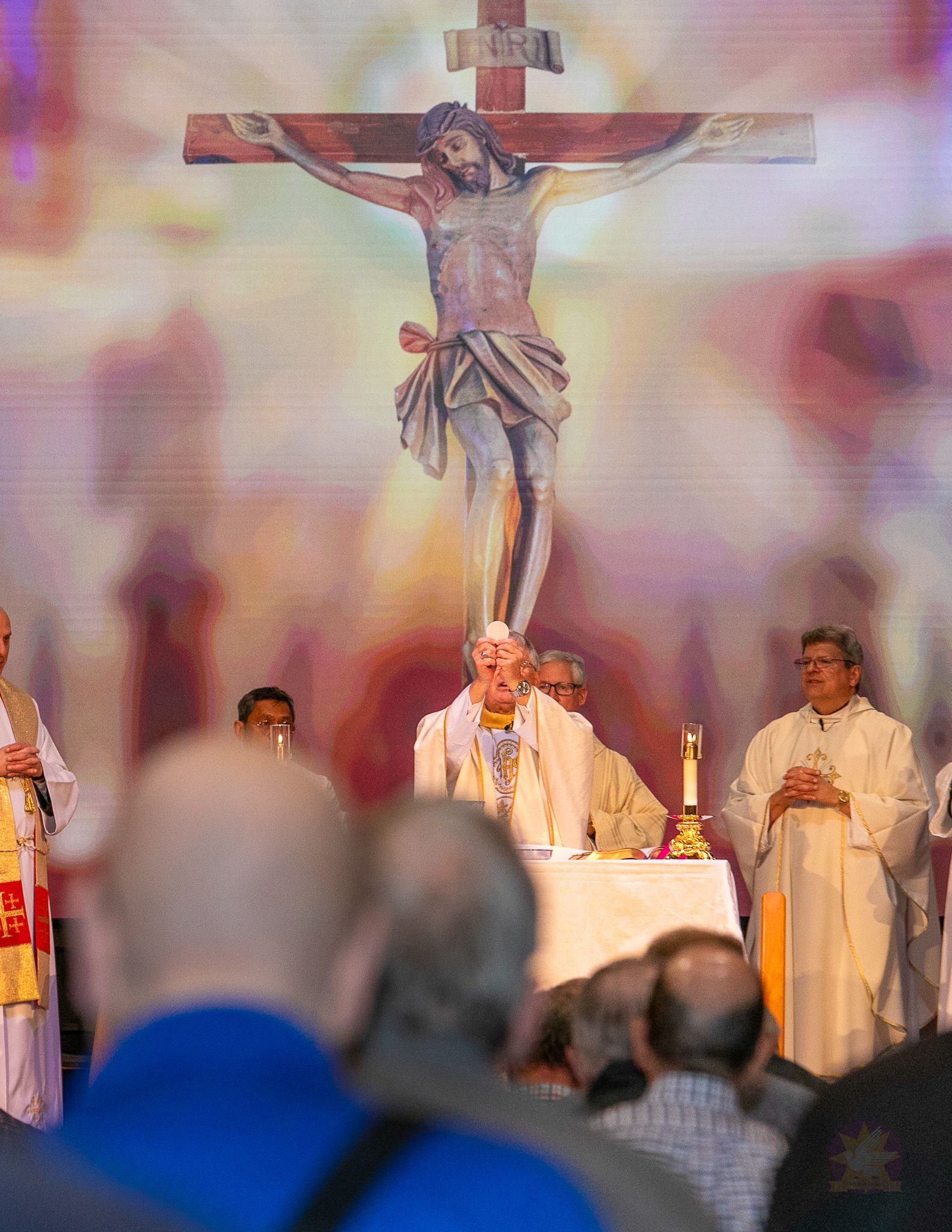 Priest at altar beneath a large crucifix during a church service, with blurred congregation in foreground