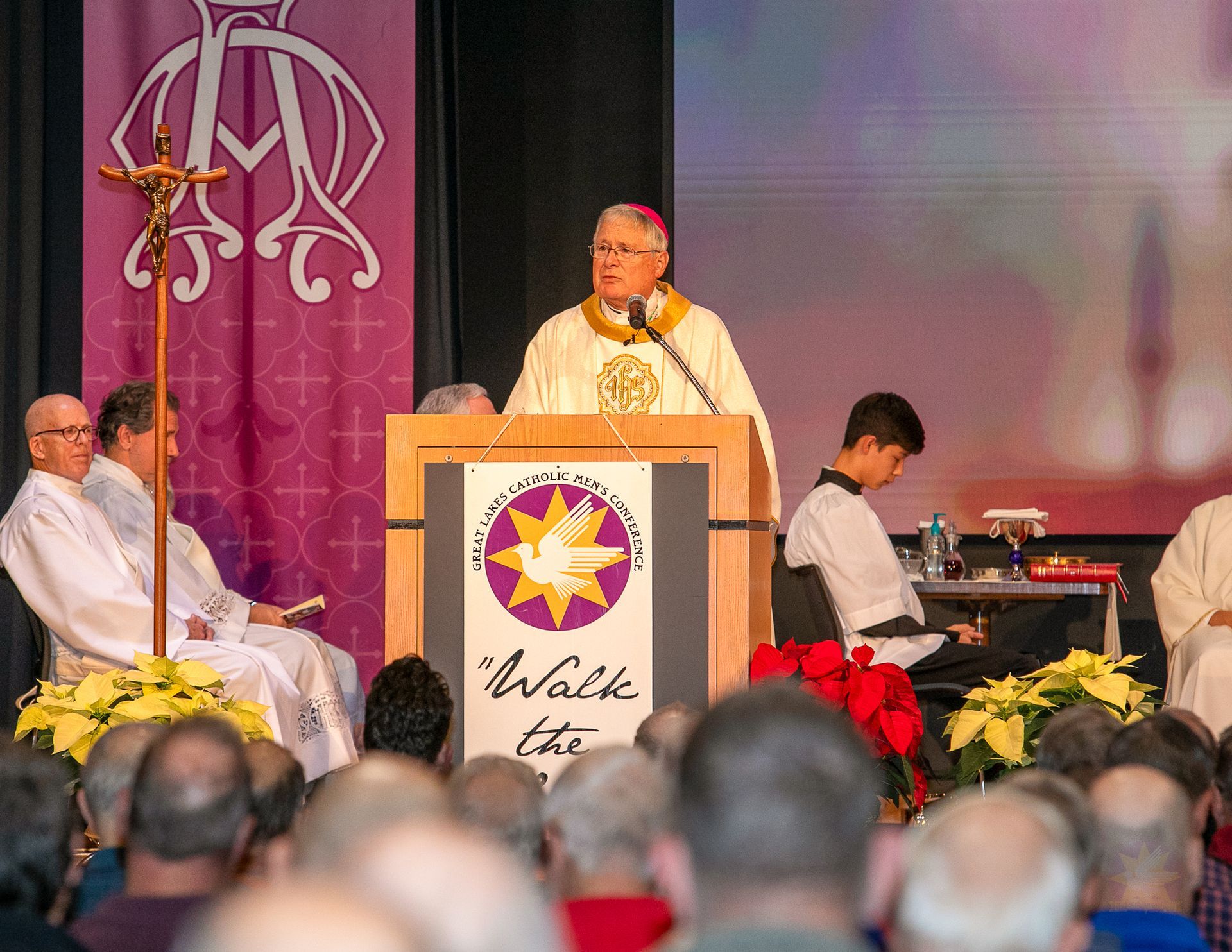 Bishop Malloy speaking at a podium during a church service, with seated participants and an audience in the foreground.