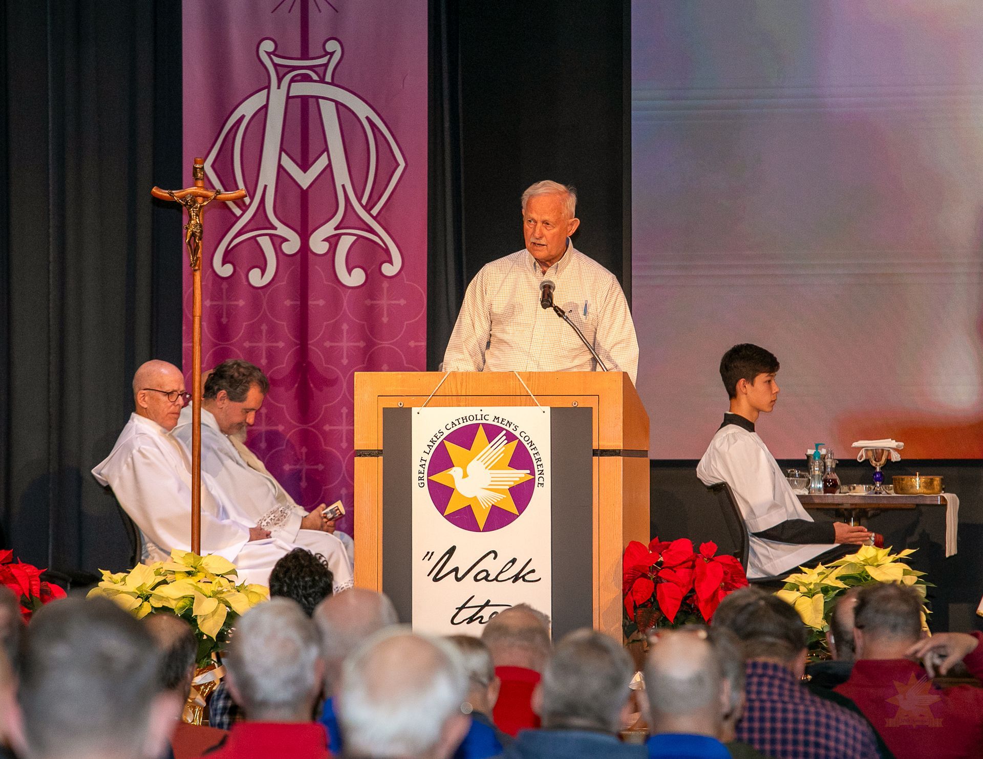 Speaker at podium on stage, flanked by seated musicians, with audience in foreground and purple backdrop behind.