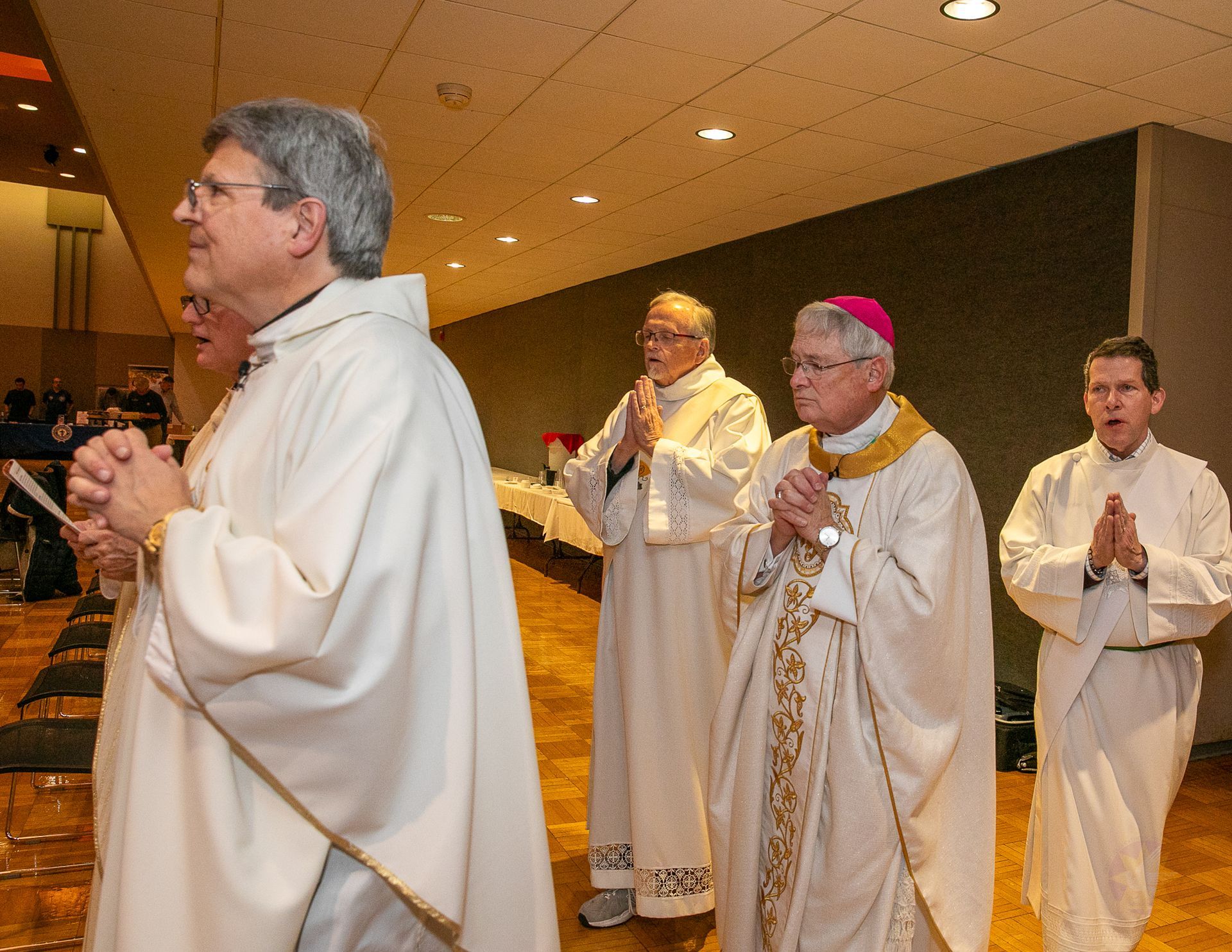Clergy in white robes walk in a procession, hands clasped in prayer.