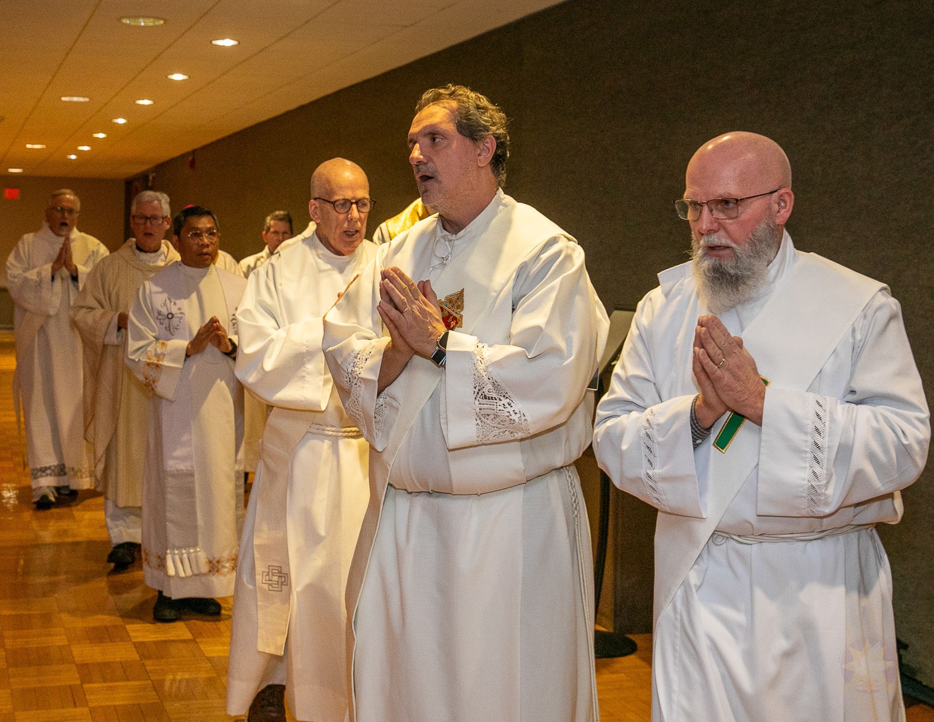 Clergy in white robes stand in a church aisle, hands clasped in prayer during a procession.