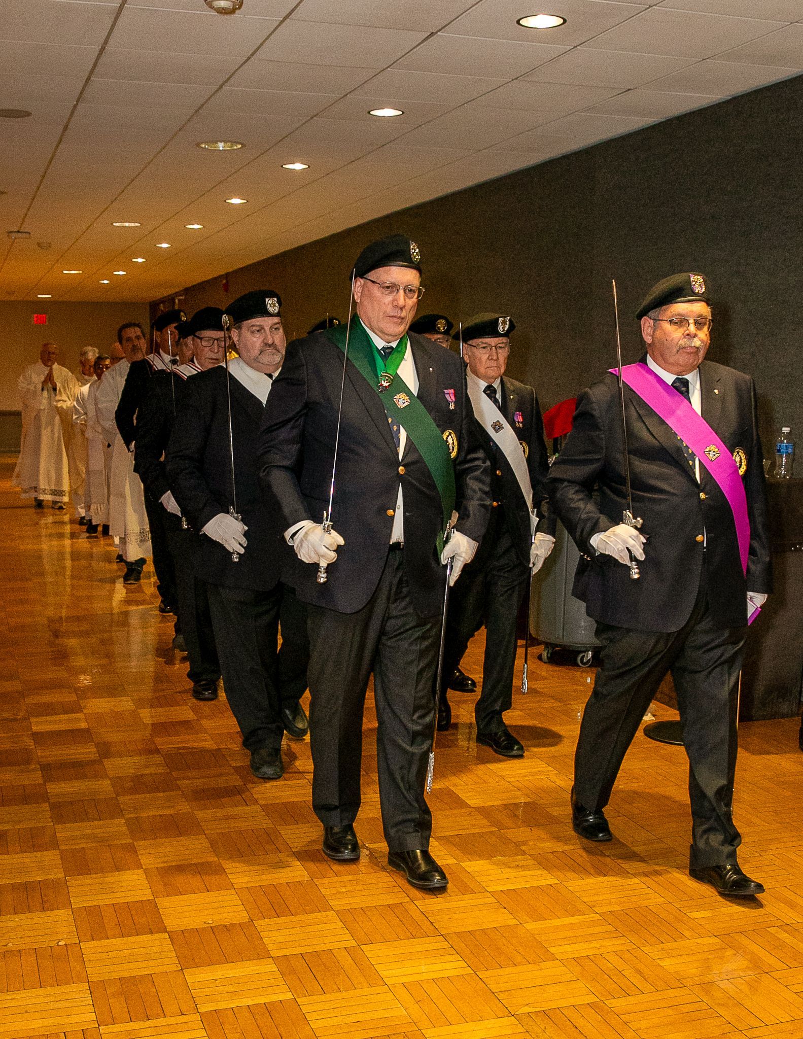 Formal procession of Knights of Columbus walking down a hallway, one wearing a green sash
