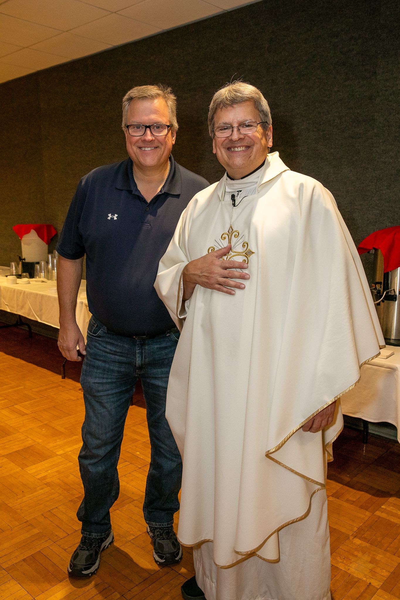 Two men standing indoors, one in a navy shirt and jeans, the other in a white robe with a cross necklace.