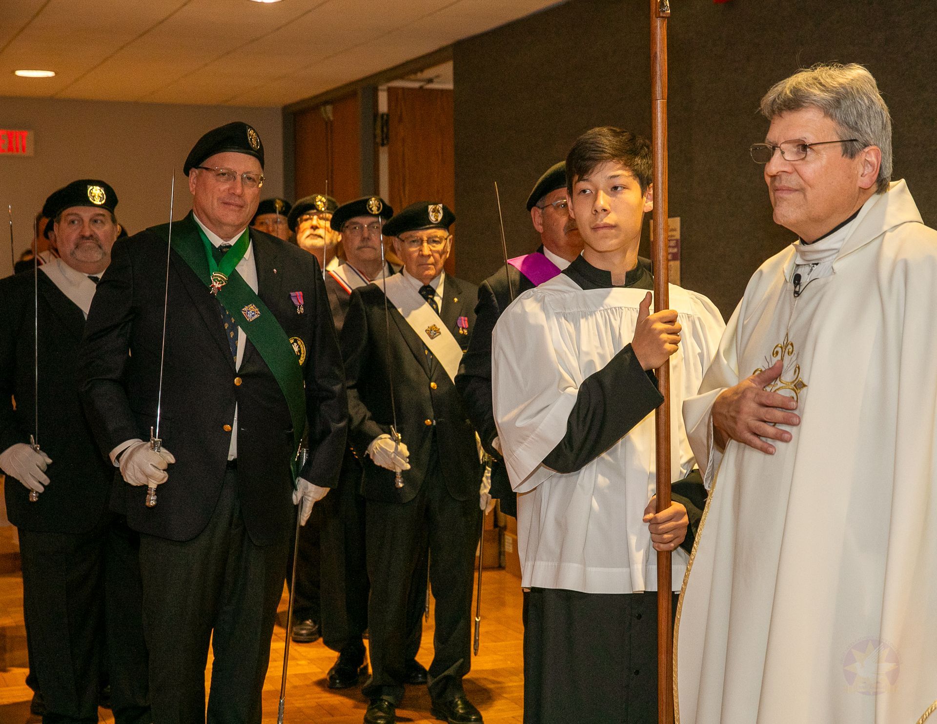 Clergy and officials standing in a hallway, one holding a processional crucifix, another in white robes.