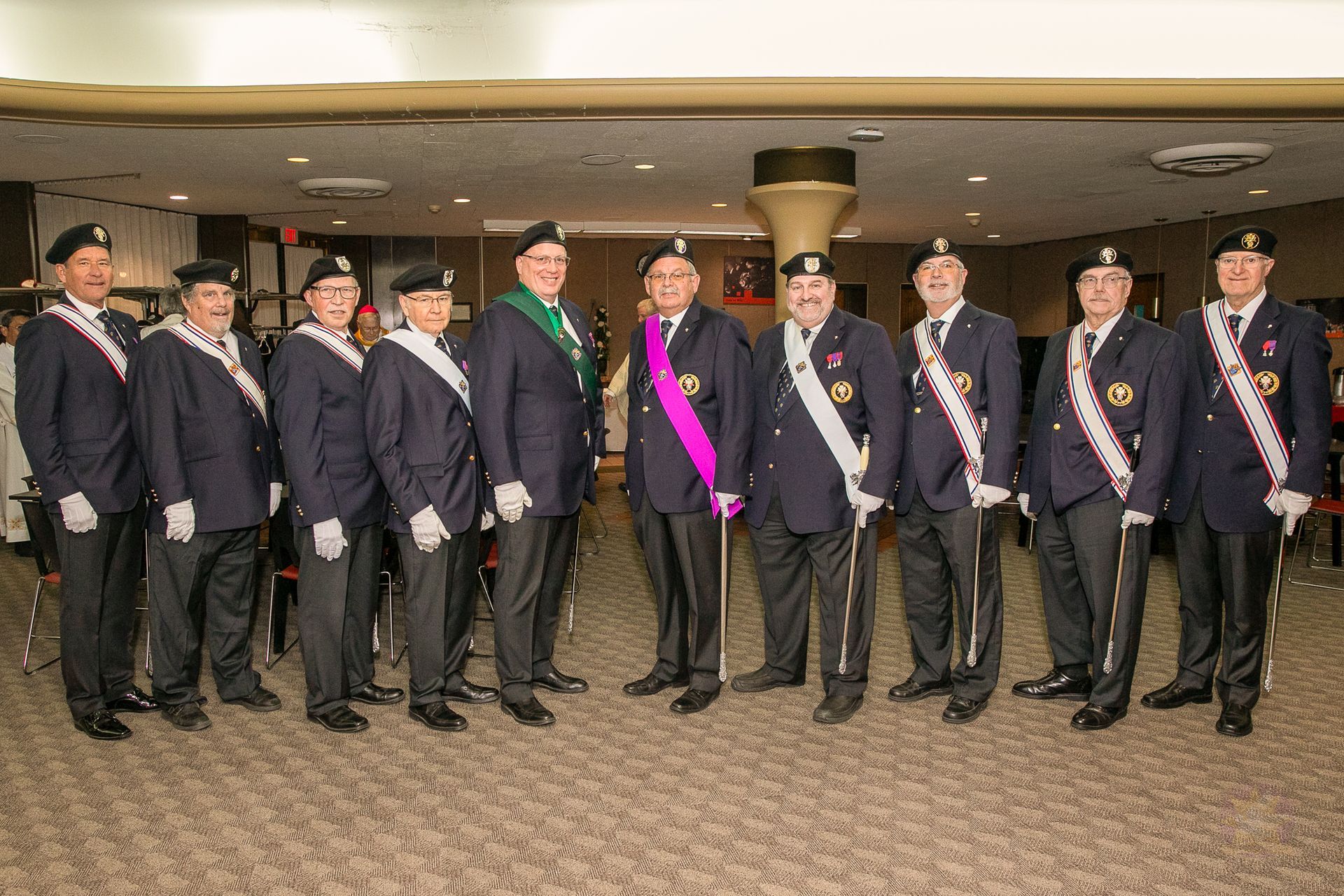 Group of formally dressed Knights of Columbus wearing sashes and medals standing in a lobby.