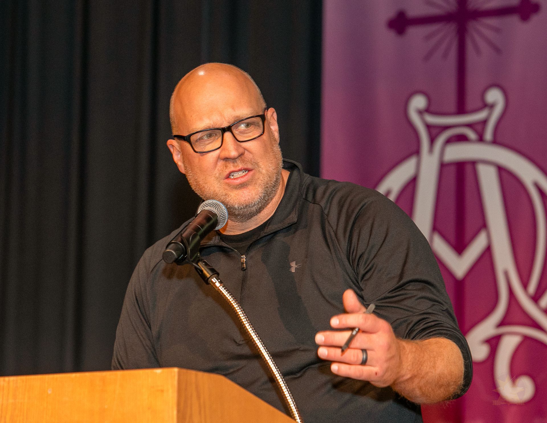 Speaker at a podium, gesturing while speaking into a microphone against a purple backdrop.